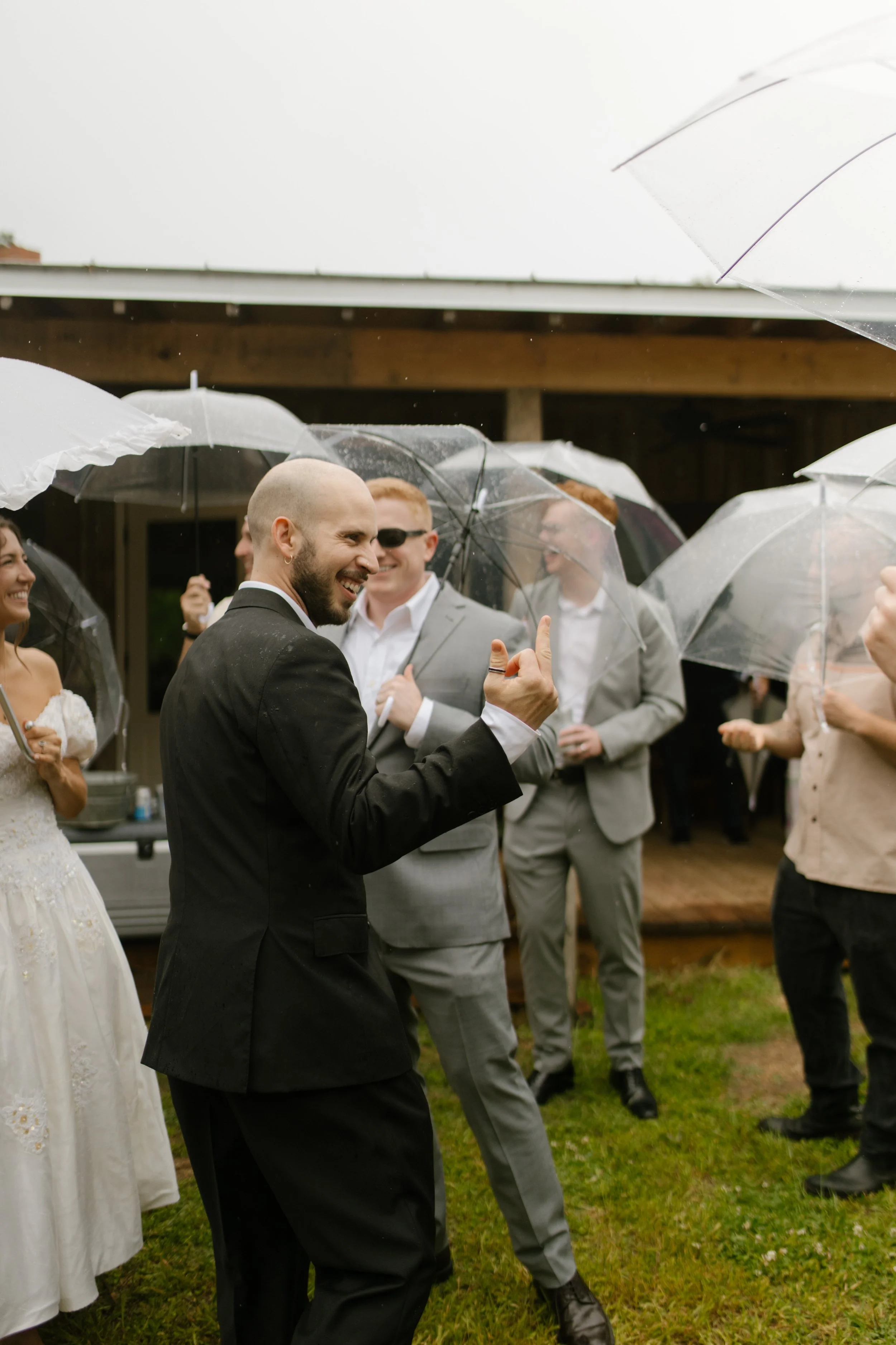 Groom smiling with friends under clear umbrellas during a rainy wedding rehearsal photography celebration.