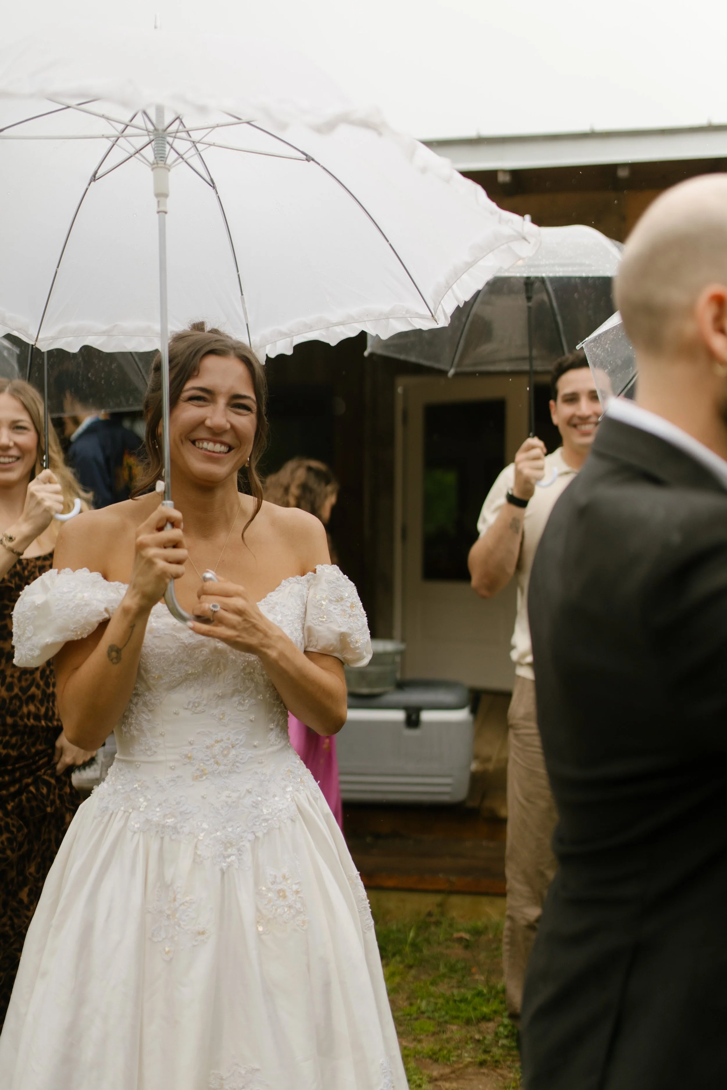 Bride smiling beneath a white umbrella as guests gather outdoors during wedding rehearsal photography in the rain.