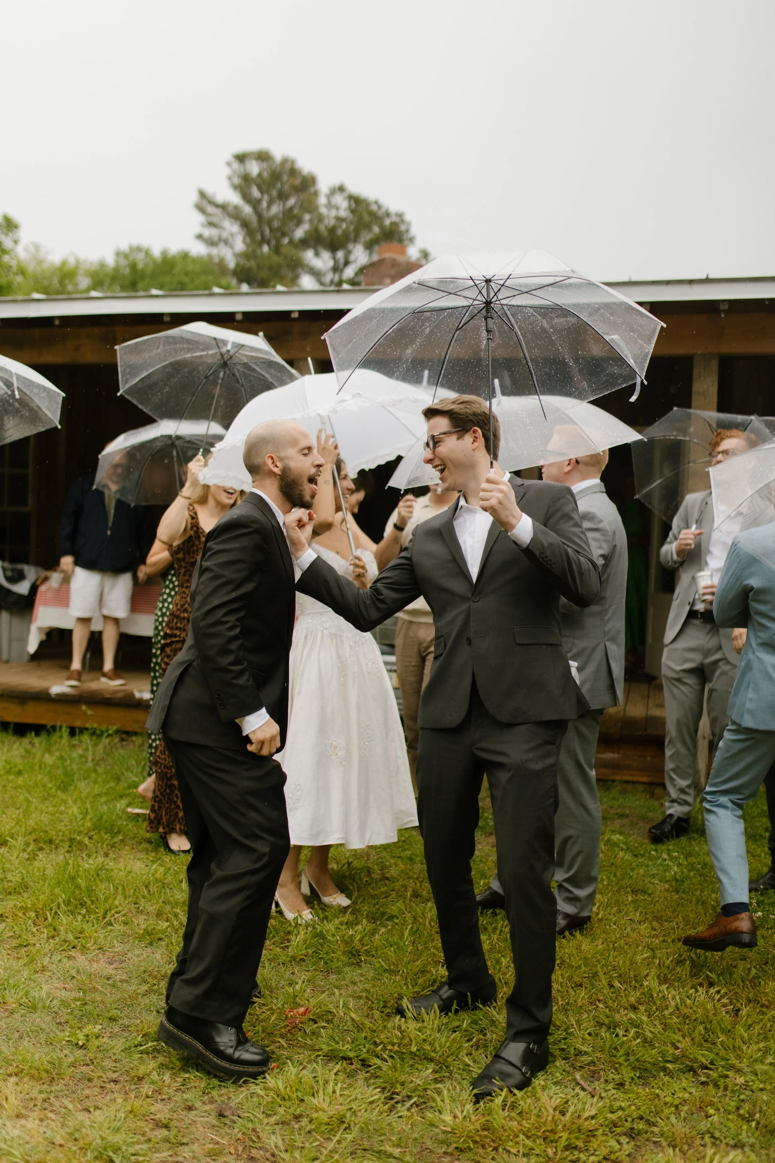 Groom laughing with a guest under clear umbrellas while friends celebrate outdoors before the wedding.