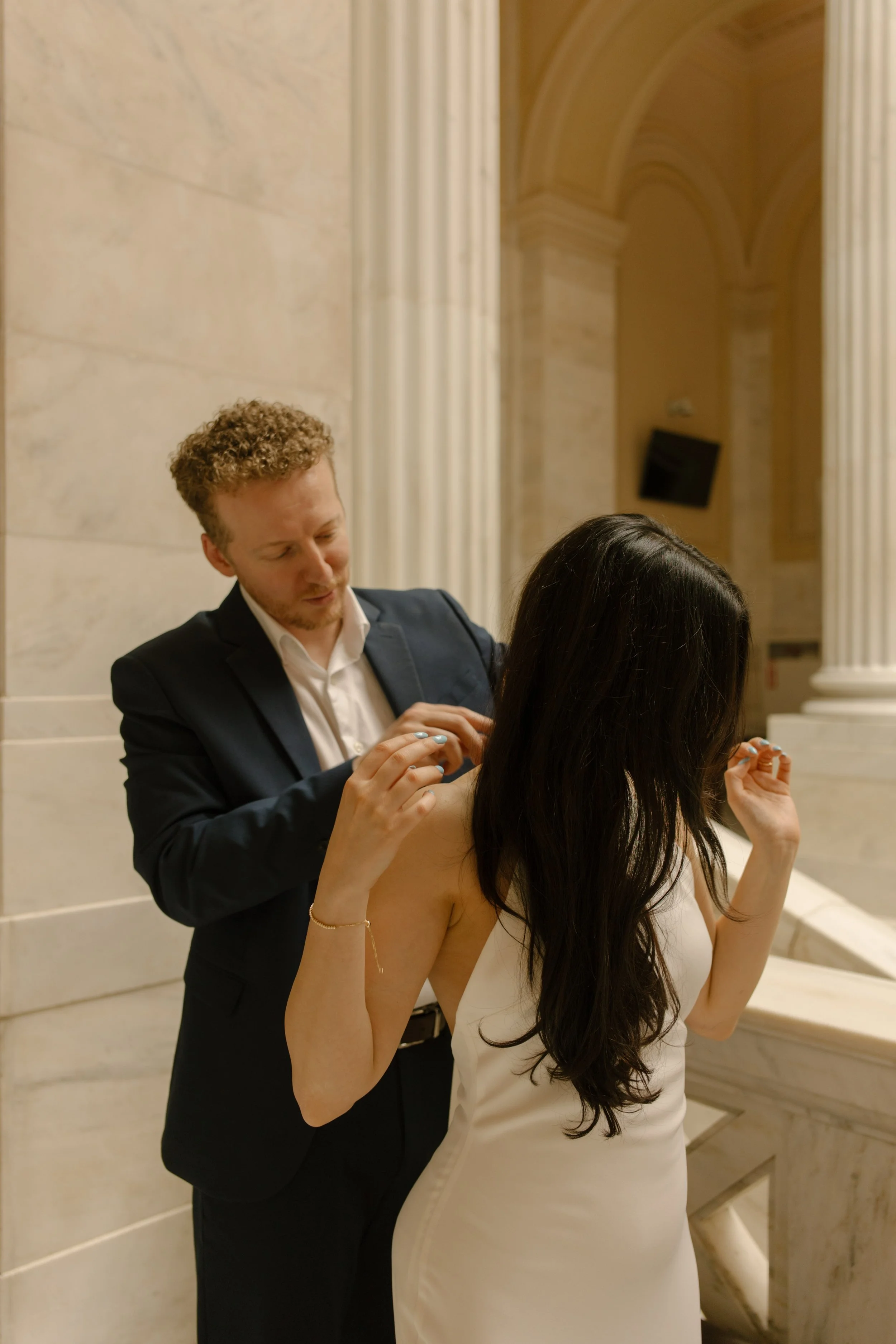 Couple shares a quiet moment while adjusting attire among elegant marble columns and stairways.