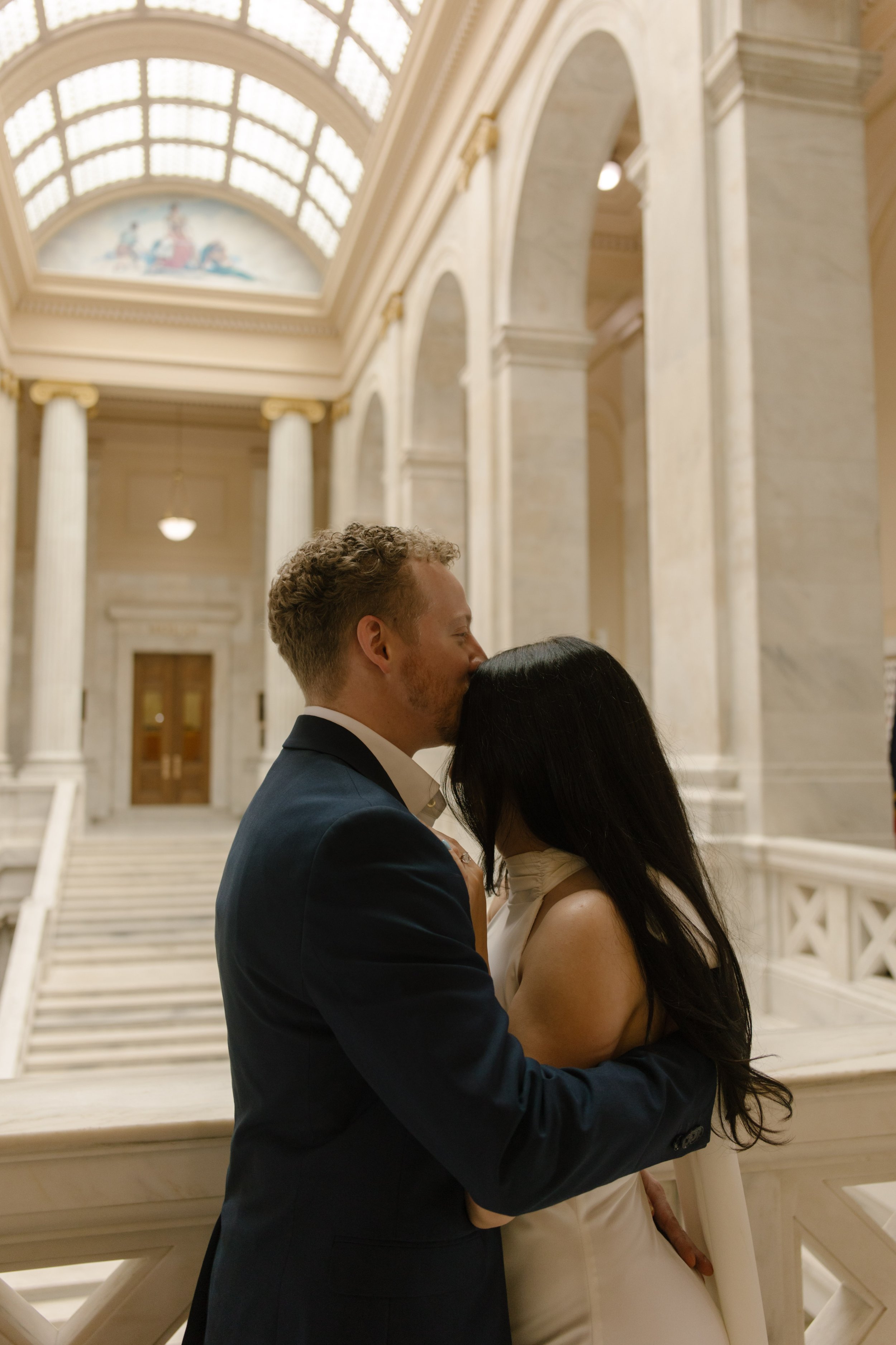 Groom kisses bride’s forehead beside ornate railings and towering columns in the courthouse atrium.