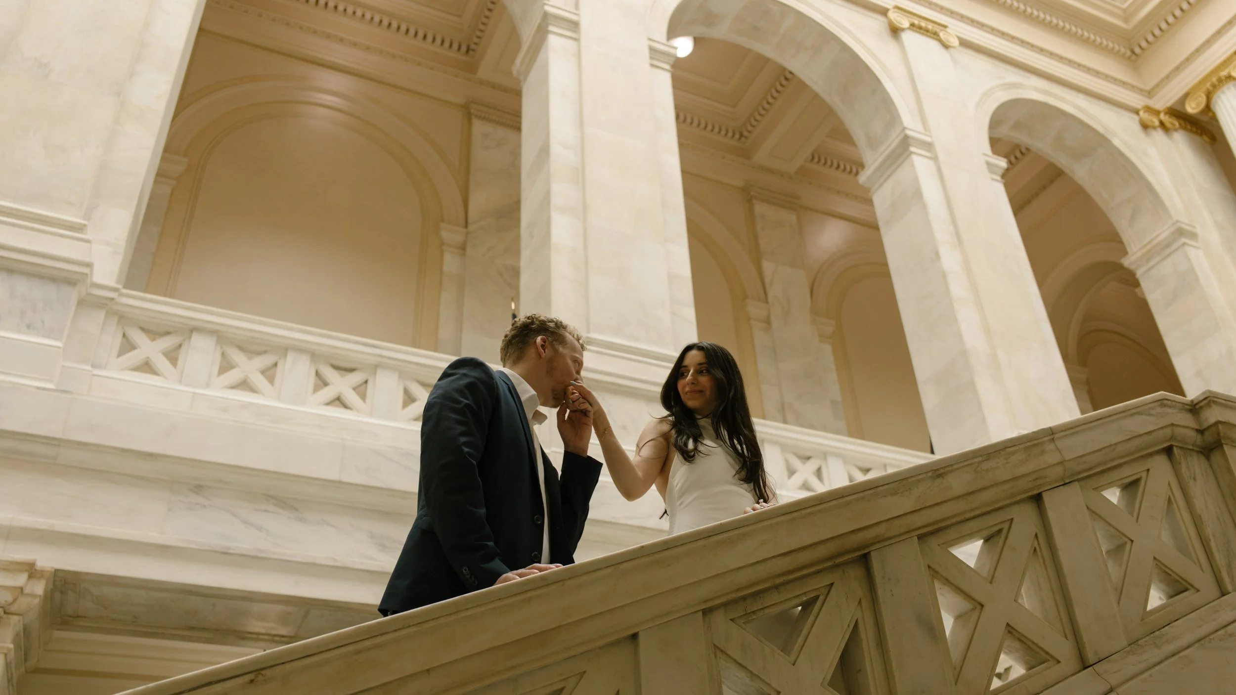Bride playfully lifts the groom’s hand for a kiss while standing on a marble staircase.