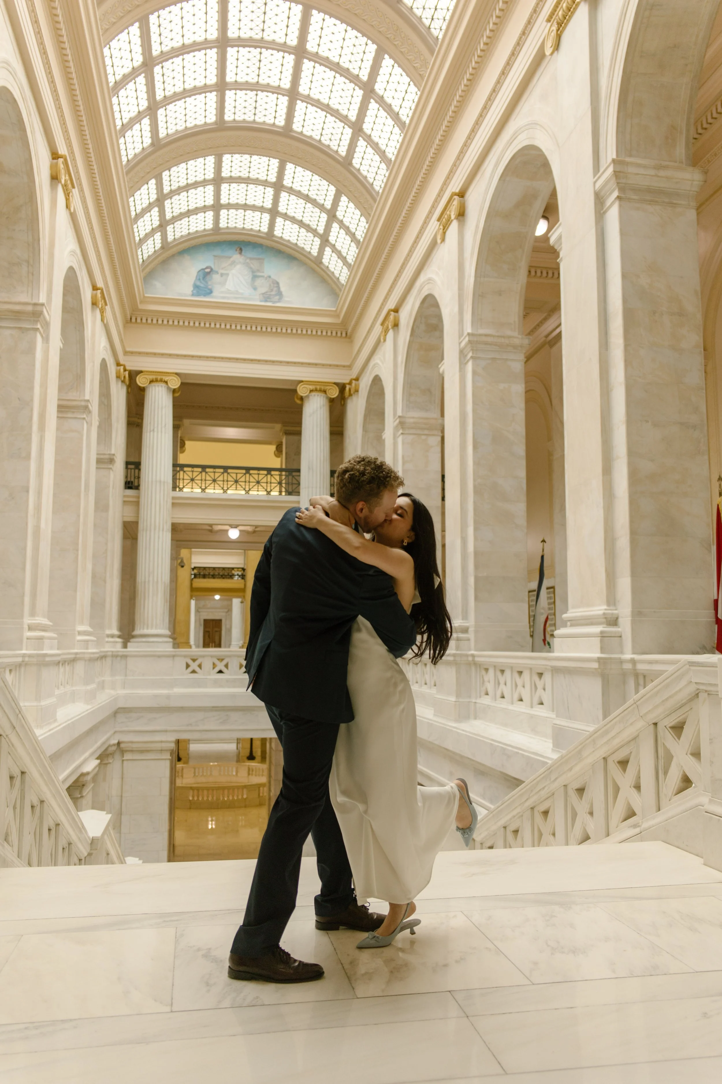 Romantic kiss on the grand marble staircase inside the Little Rock courthouse beneath soaring arches and skylights.