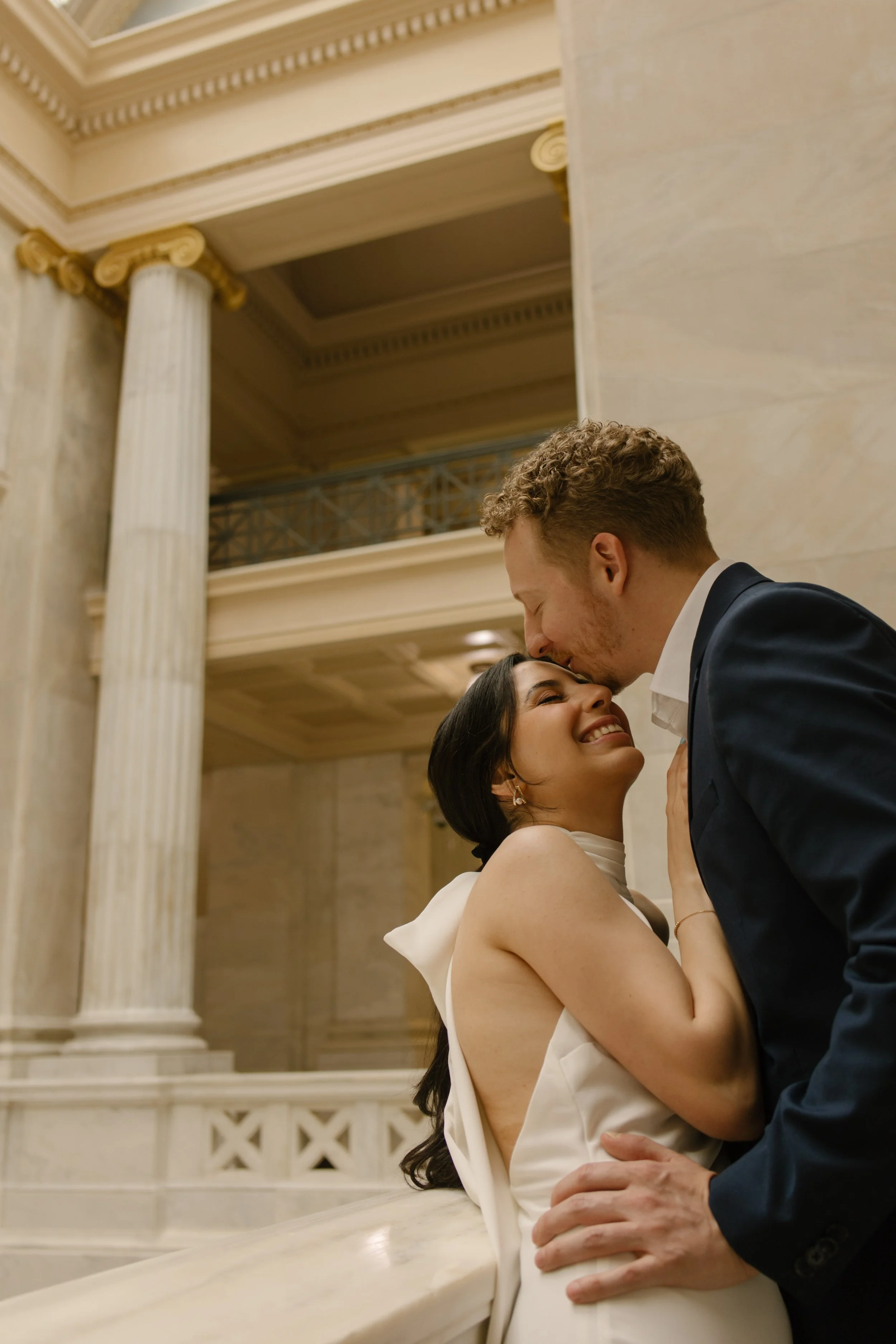 Groom kisses smiling bride beside marble columns during a romantic Little Rock courthouse photoshoot.