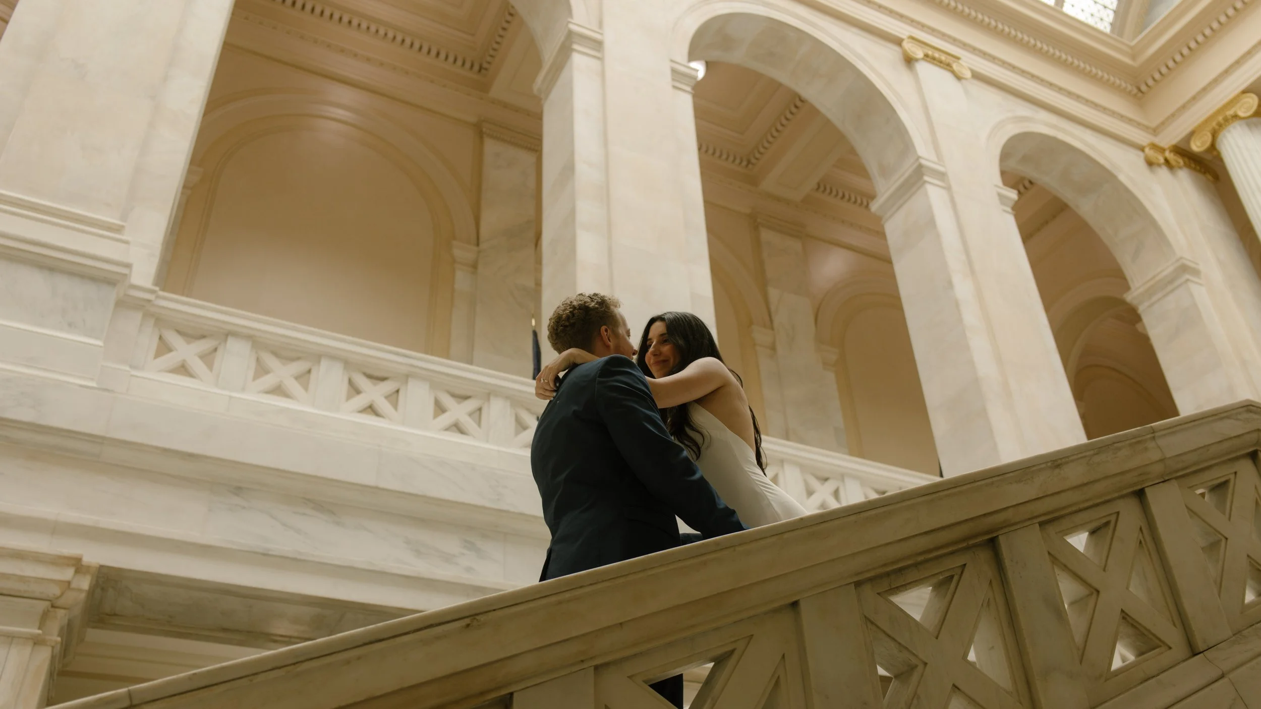 Couple embraces on a marble staircase surrounded by classic courthouse architecture and arched hallways.