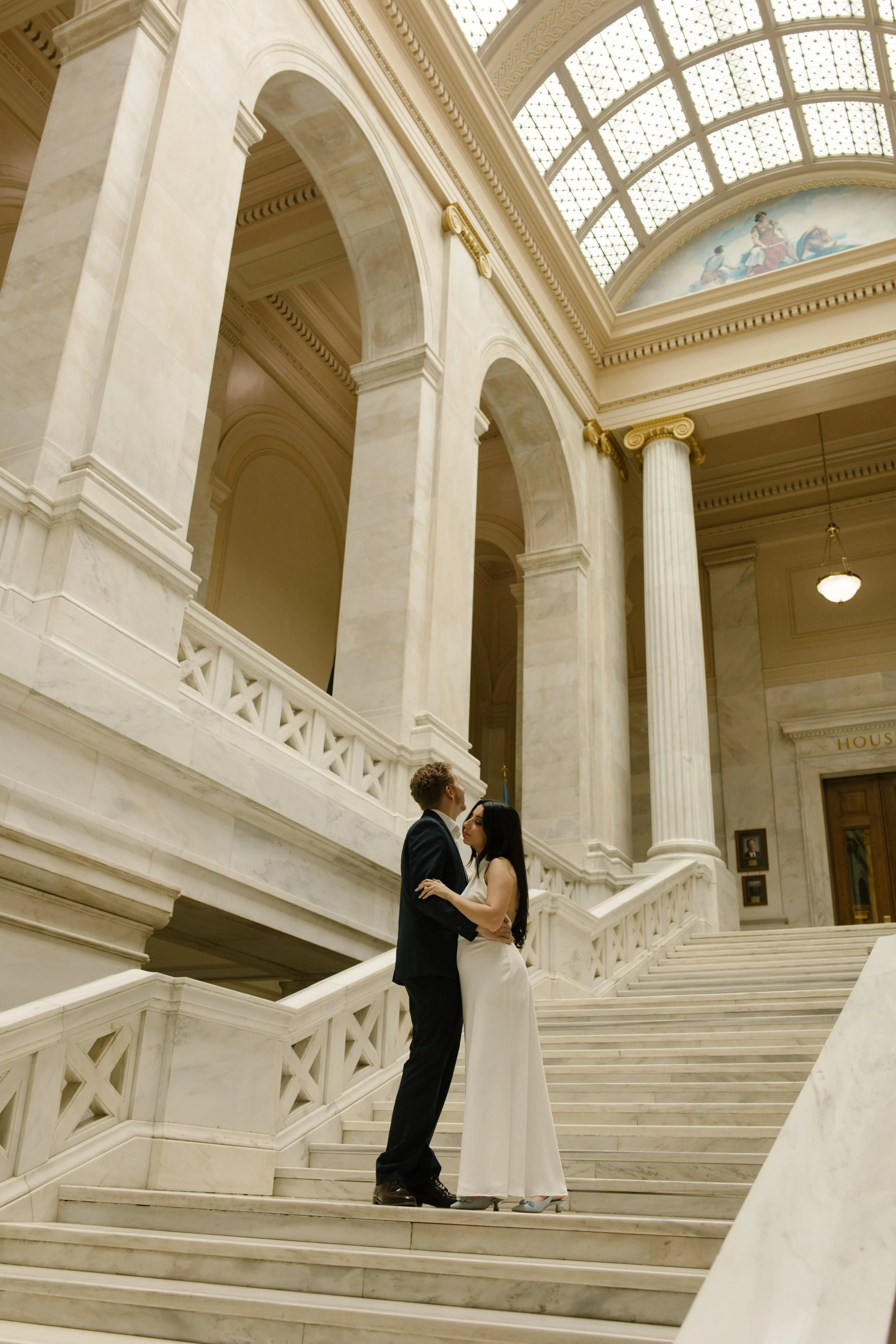 Couple embraces on the grand staircase inside the Little Rock courthouse beneath soaring marble arches and a glass skylight.