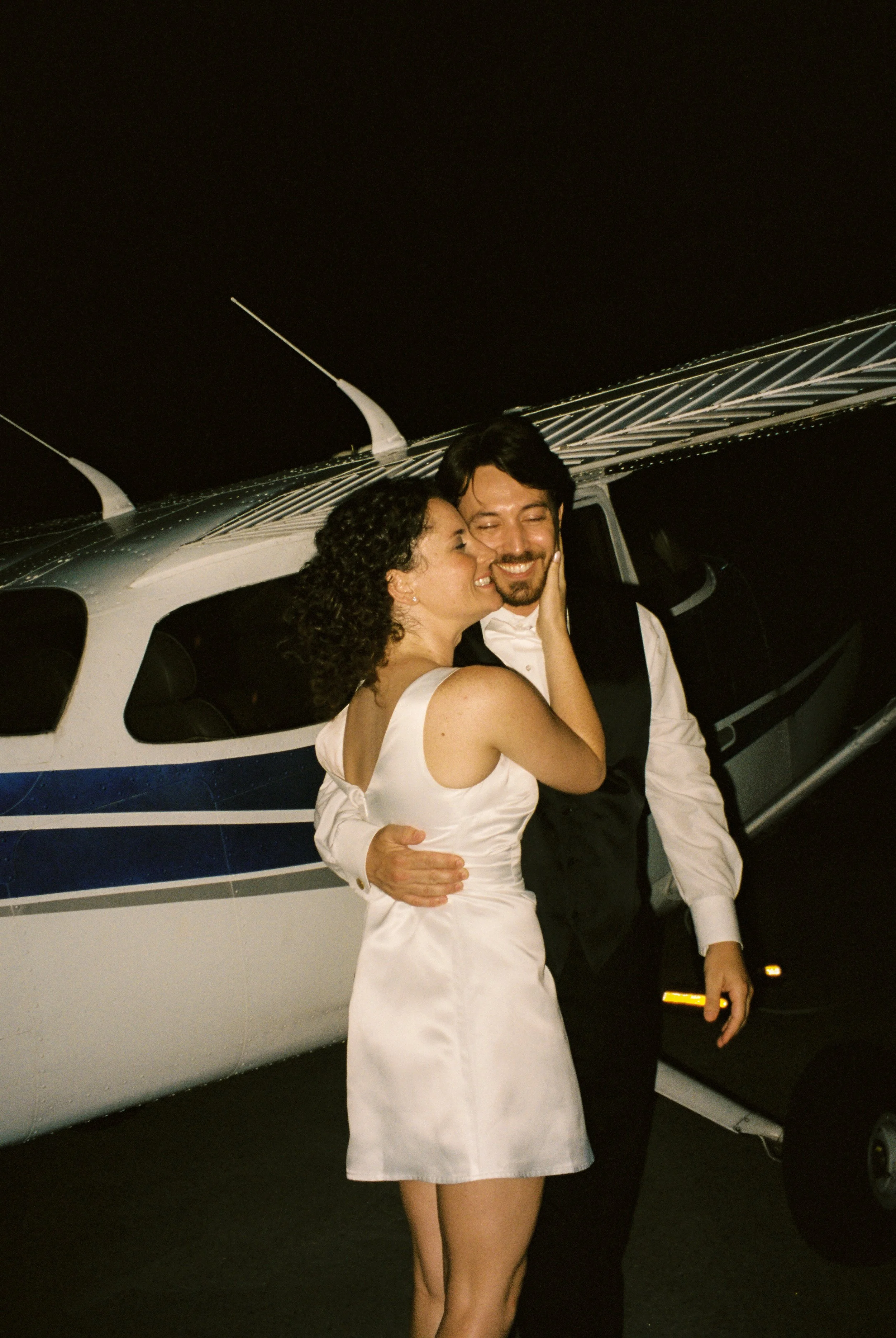 Newlywed couple embraces beside a small airplane after their wedding celebration in a candid documentary wedding photography image.