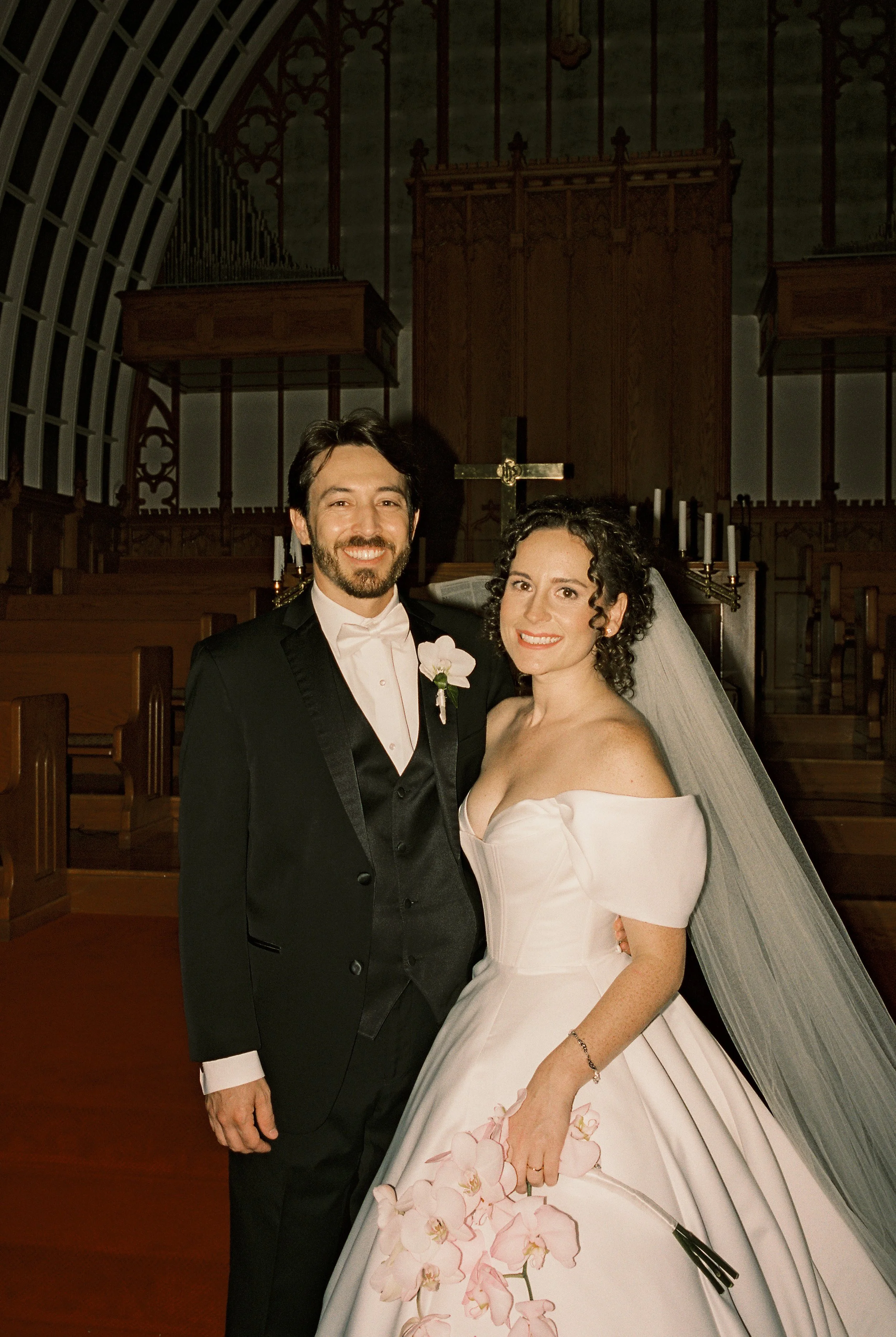 Bride and groom pose together at the church altar after their ceremony, holding orchids and smiling at the camera.