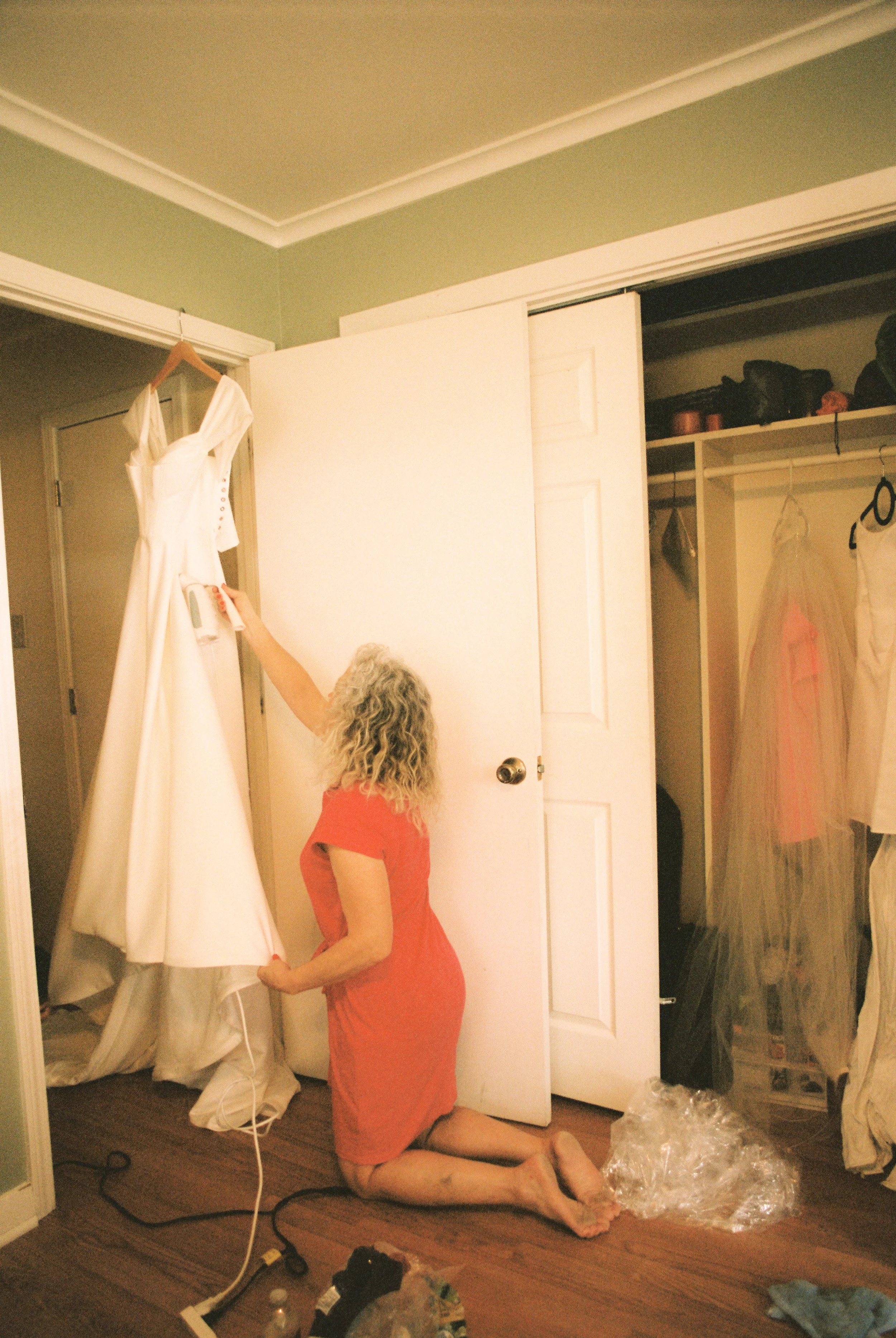 Woman steams a wedding dress hanging in a bedroom before the ceremony, captured through documentary wedding photography.