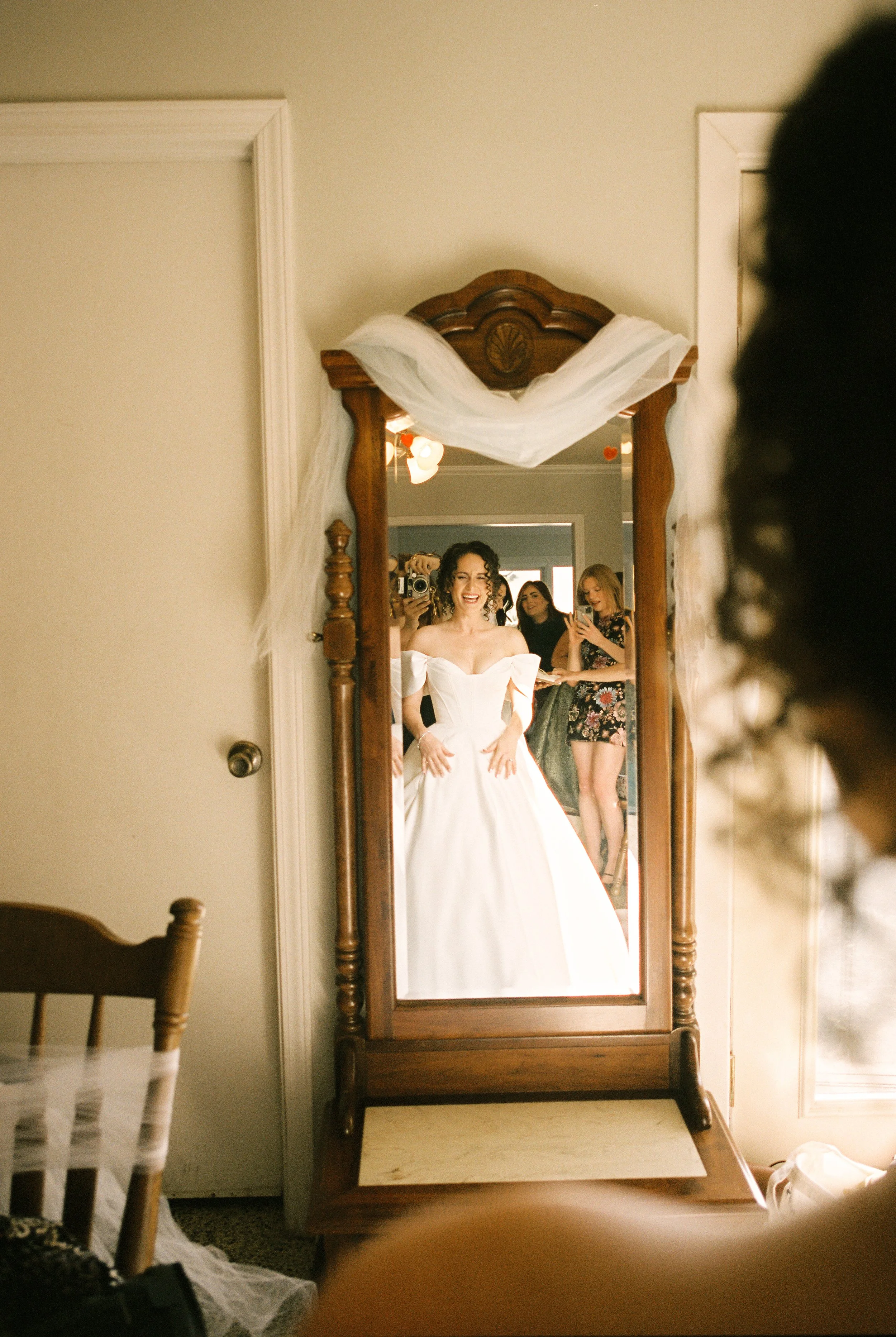 Bride sees herself in her wedding gown reflected in a vintage mirror while friends watch during wedding preparations.