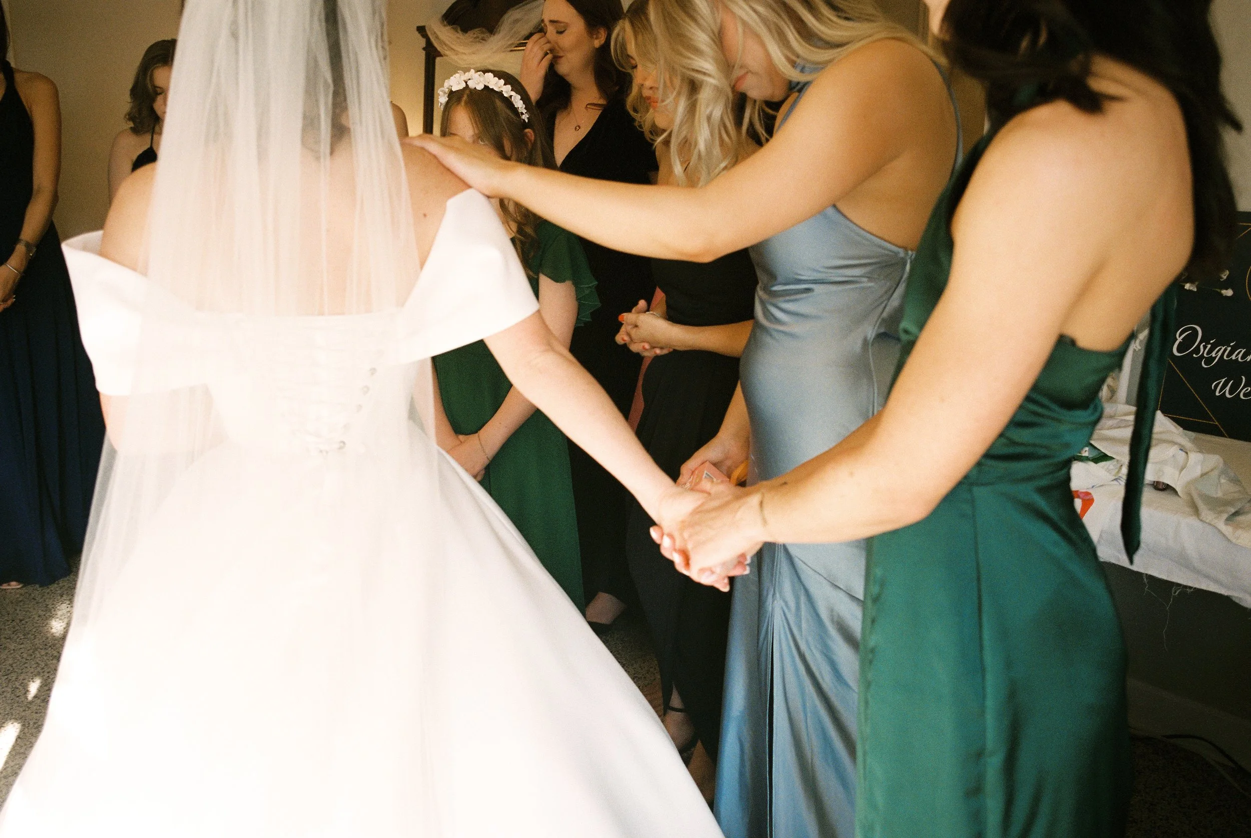 Bride and bridesmaids hold hands in a prayer circle before the wedding ceremony in a documentary wedding photography moment.