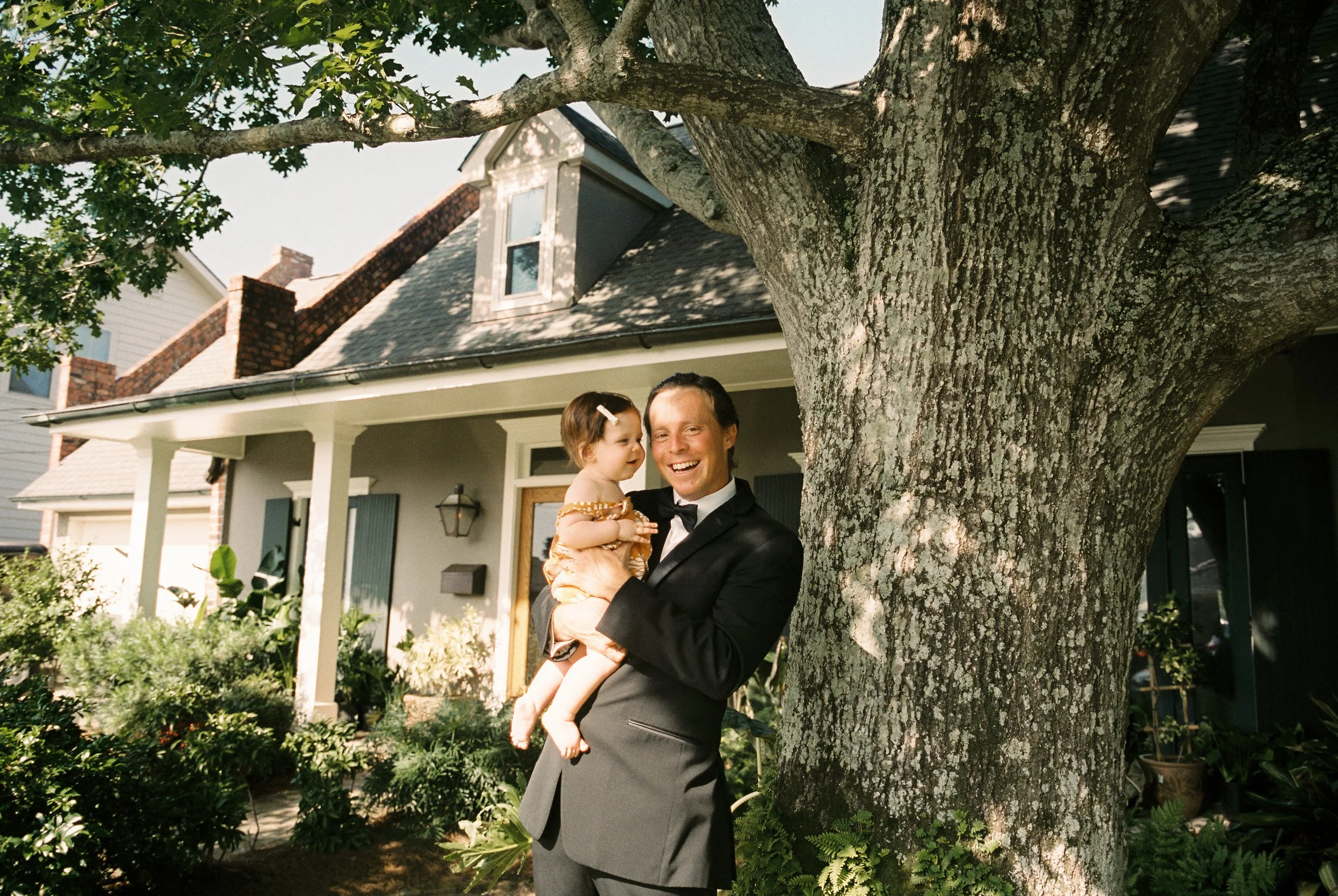 Family member holds a child beneath a large tree outside a historic home on the wedding day.