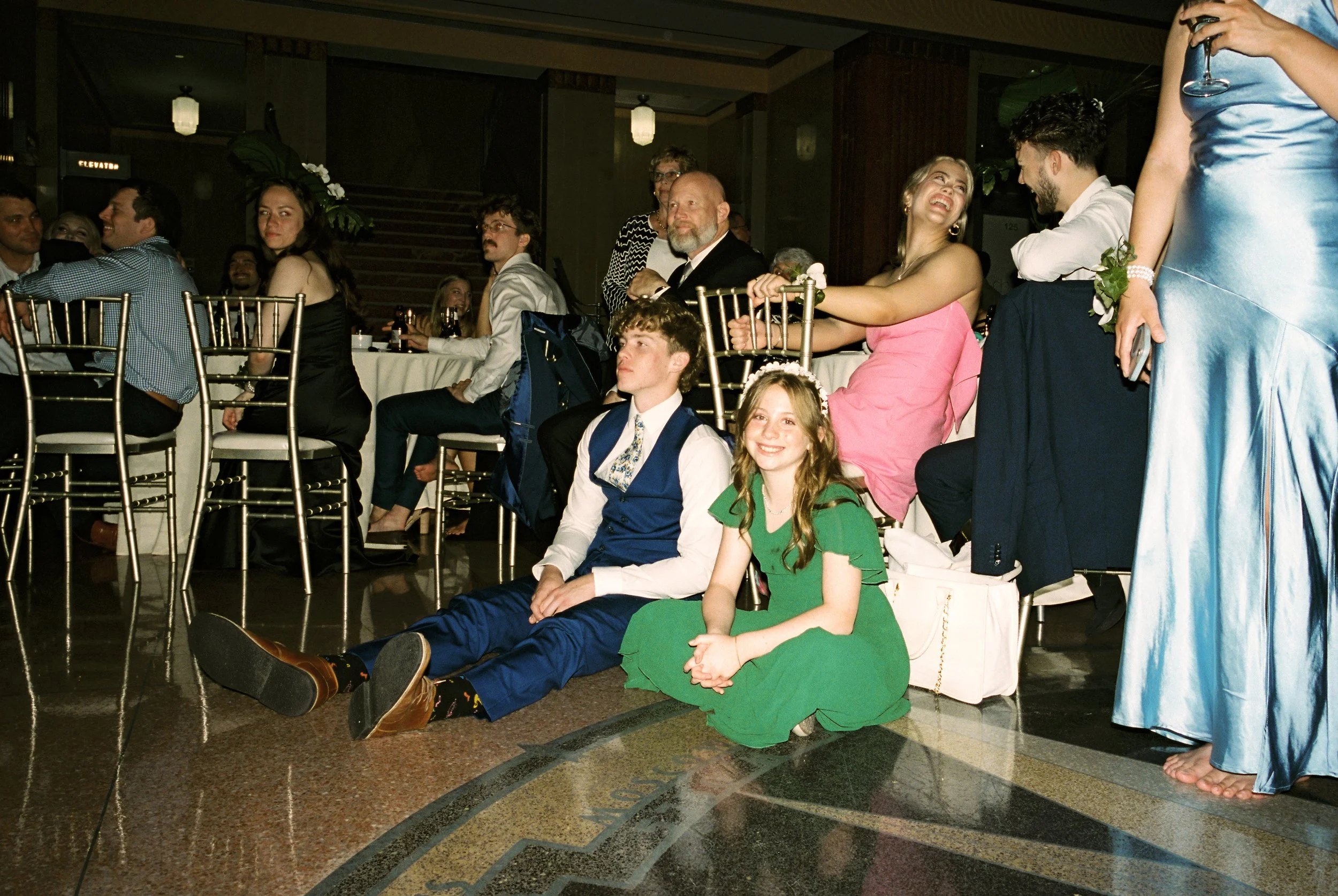 Wedding guests, including children seated on the floor, watch the reception festivities in a candid documentary wedding photography moment.