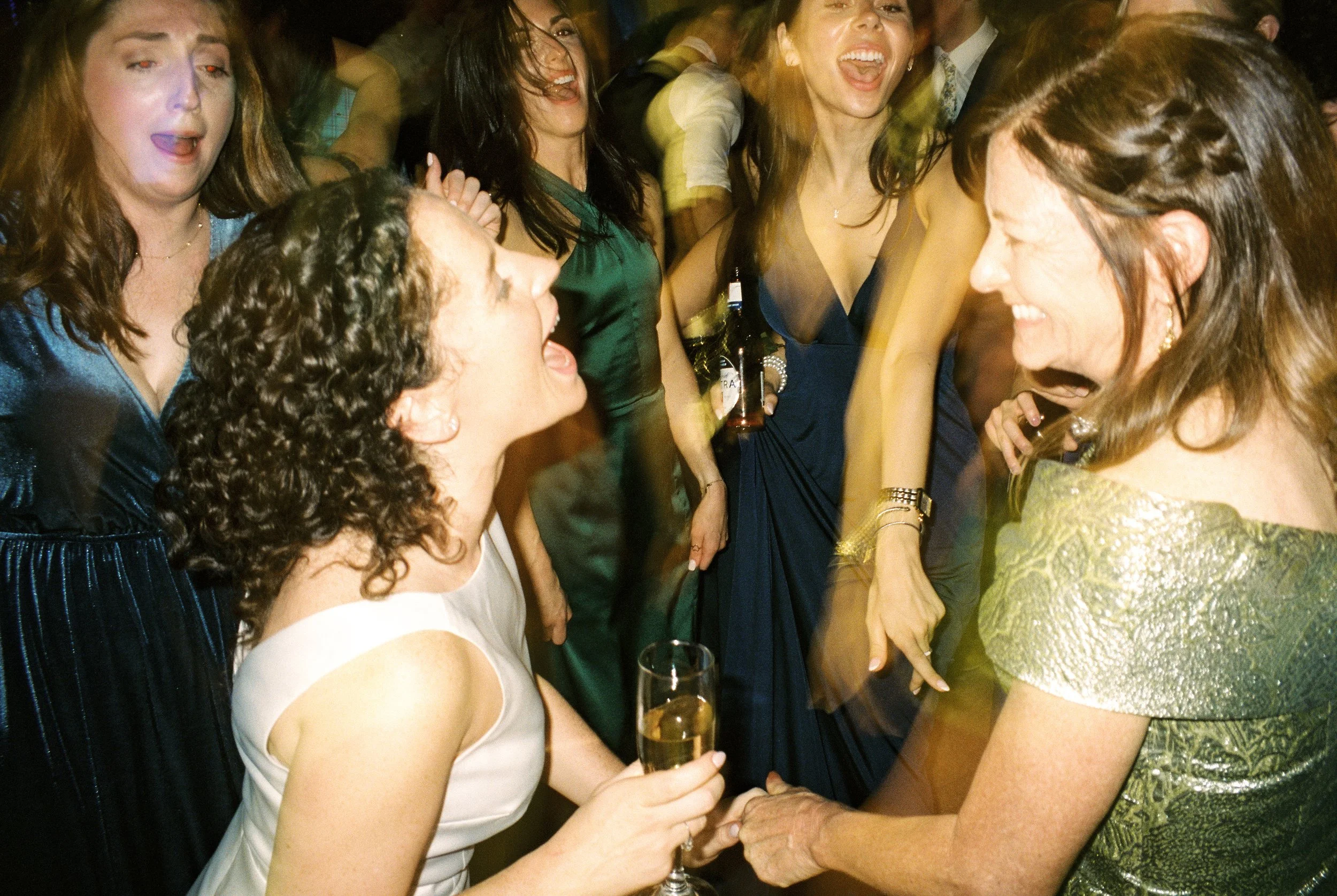 Bride dances with guests on a crowded dance floor, holding a champagne flute during an energetic reception moment.