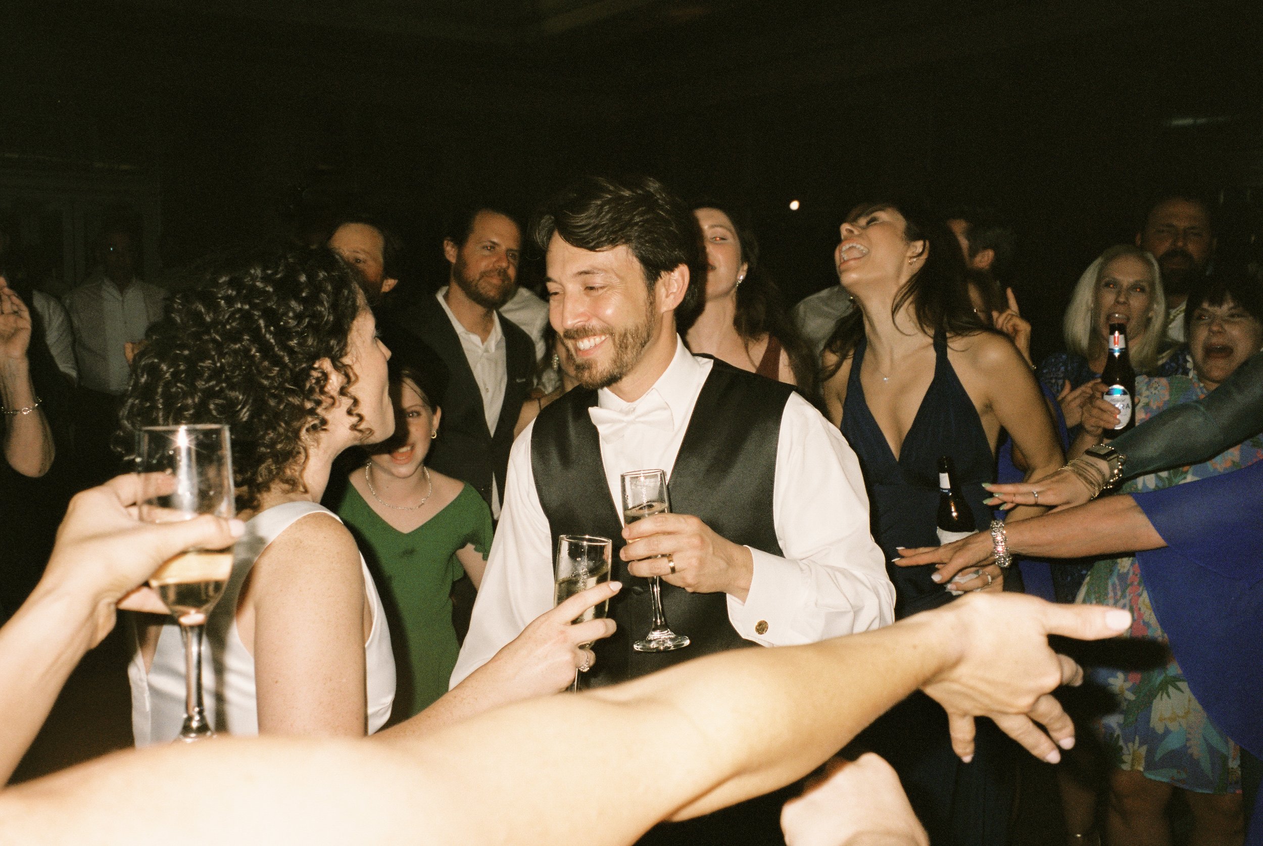 Bride and groom celebrate on the dance floor surrounded by cheering guests raising glasses and hands in the air.