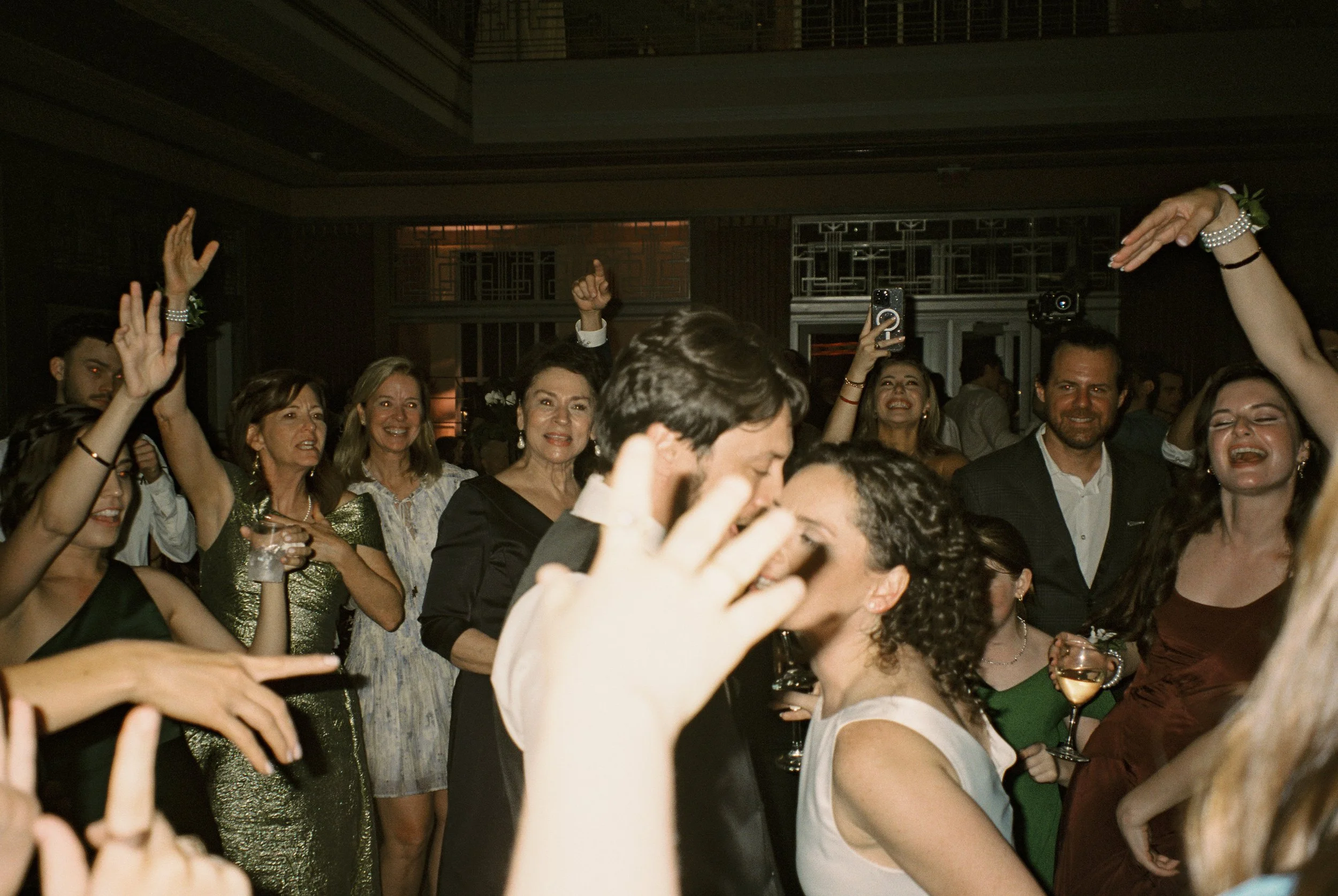 Bride and groom celebrate on the dance floor surrounded by cheering guests raising glasses and hands in the air.