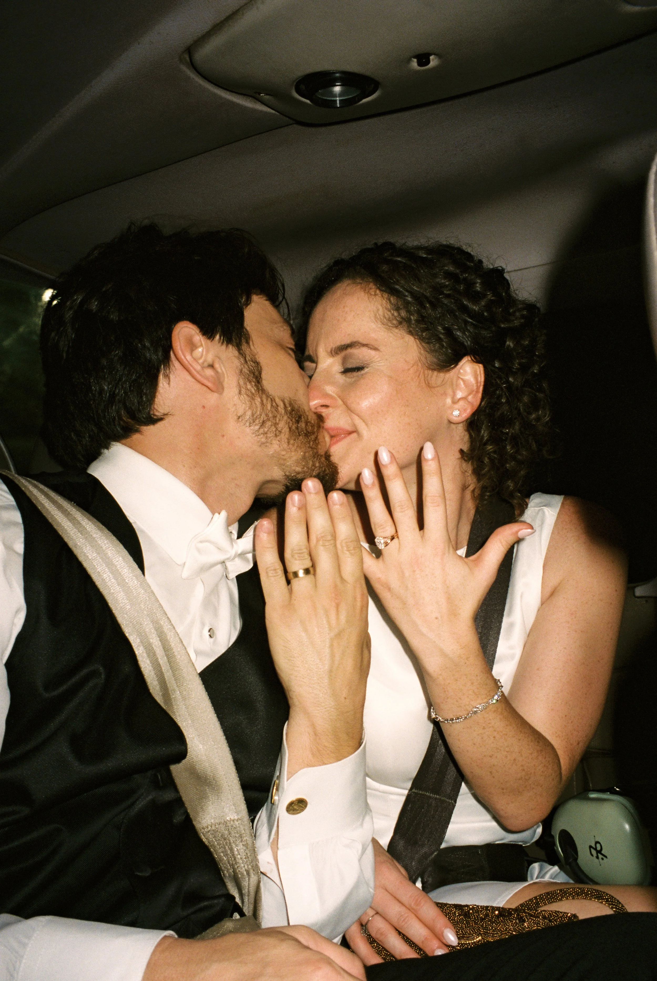 Bride and groom sitting side-by-side in a vehicle after the reception, sharing a kiss while showing their wedding rings to the camera.