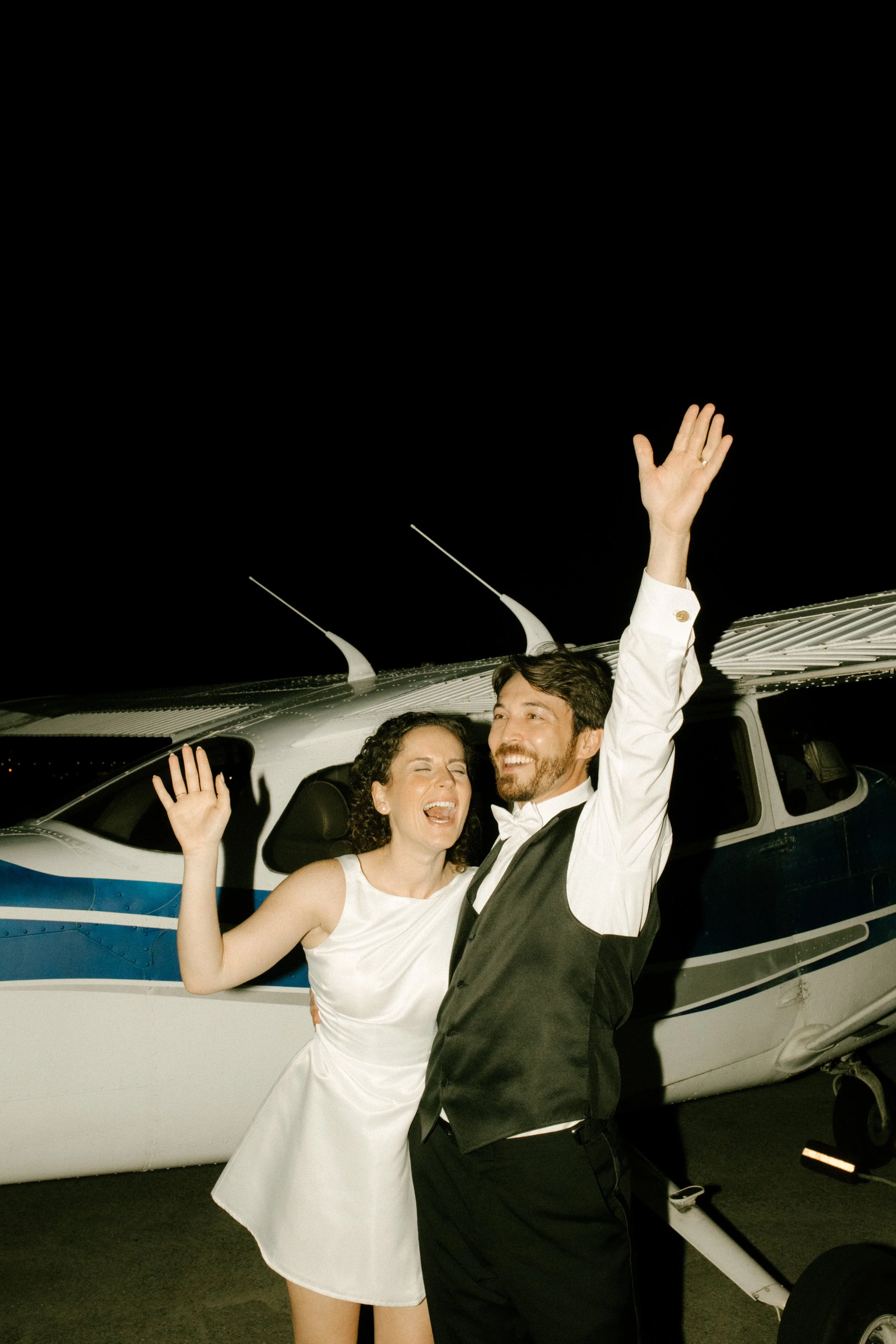 Couple celebrating beside a small airplane at night, waving enthusiastically with arms raised.
