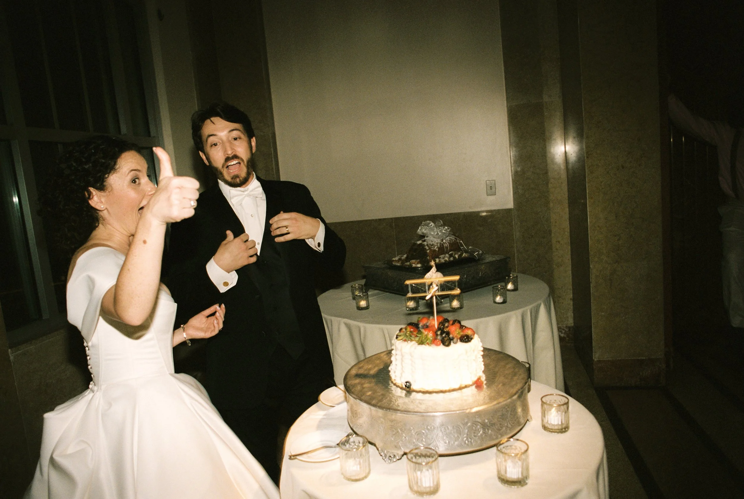 Bride gives a thumbs-up beside the wedding cake while the groom reacts playfully in the background.