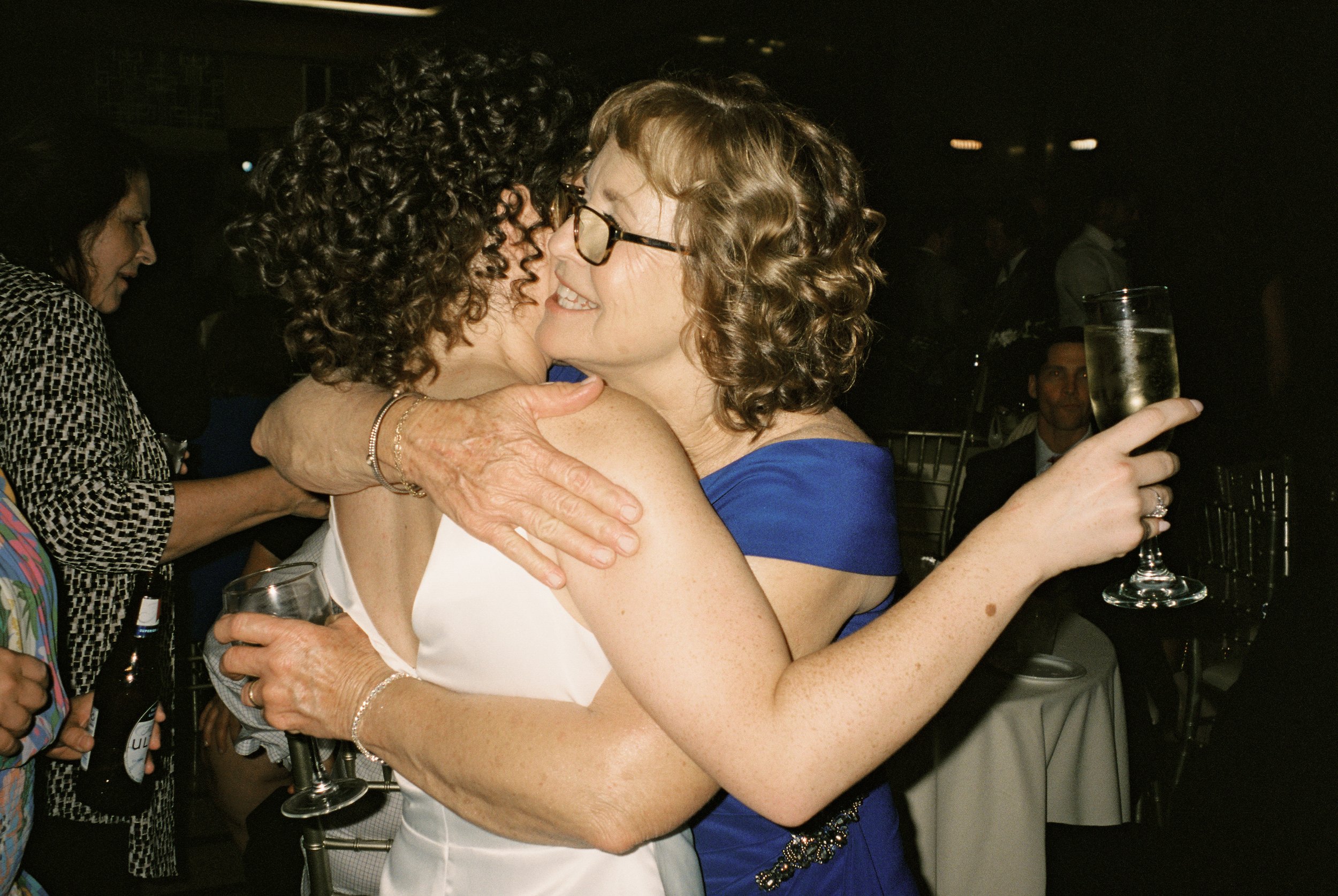 Warm embrace between the bride and an older woman in a blue dress, each holding a champagne flute.