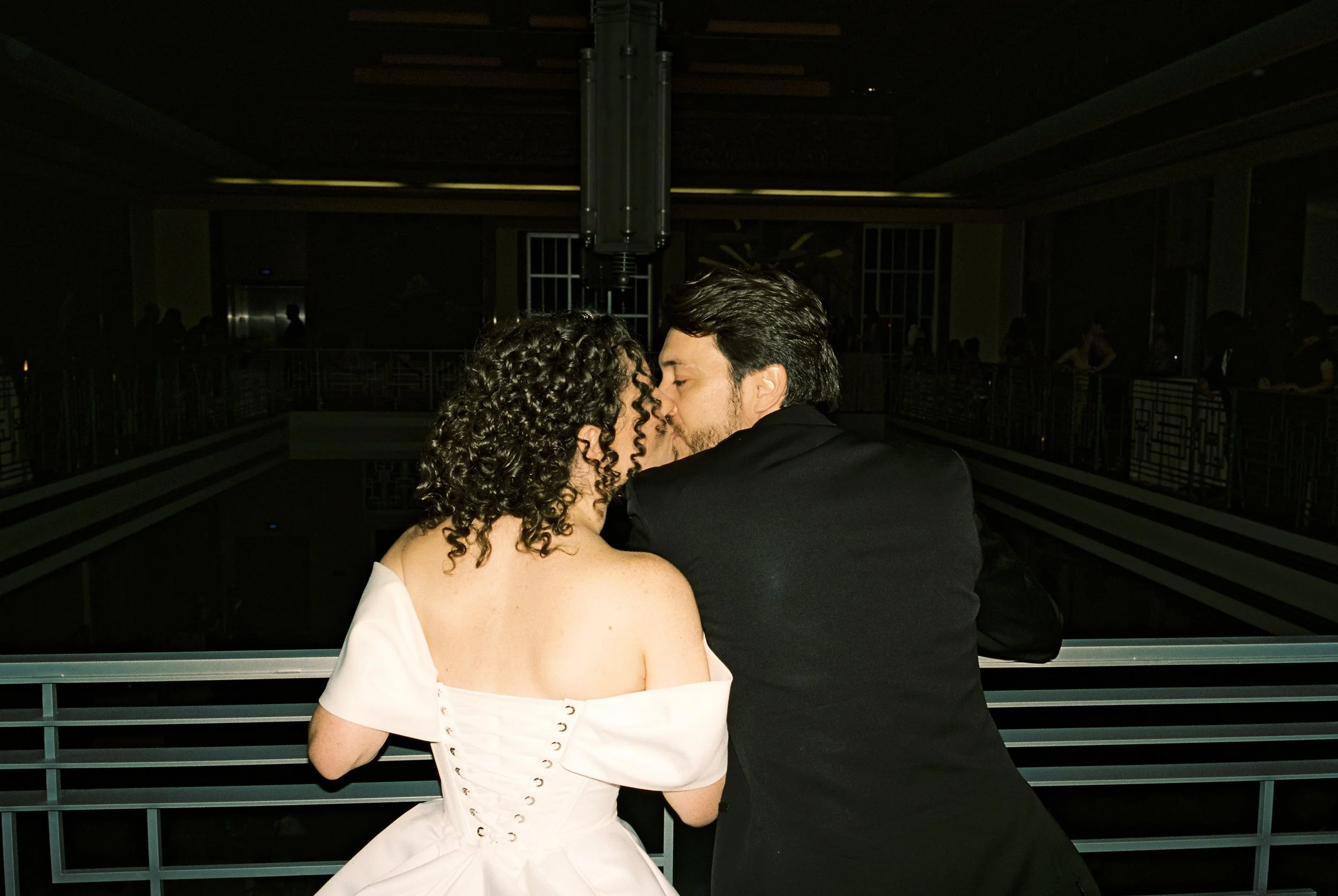 Couple leaning over a balcony railing, sharing a kiss in a dimly lit reception venue.