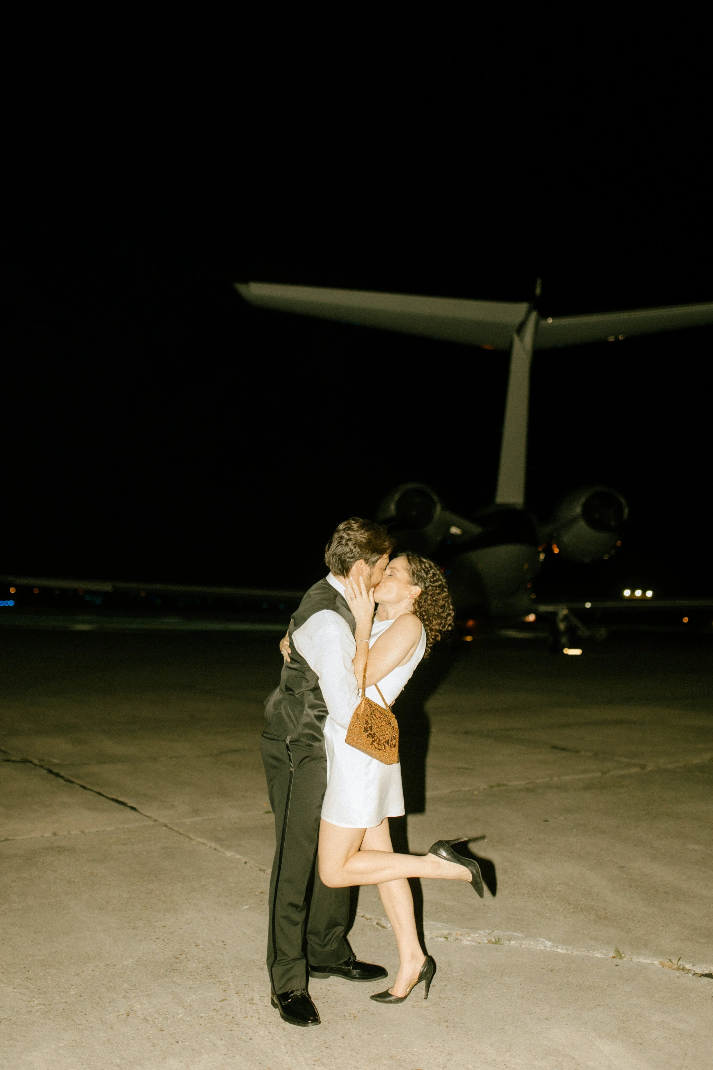 Couple kissing on an airport tarmac at night, with a large aircraft visible in the background, creating a cinematic ending to the wedding celebration.