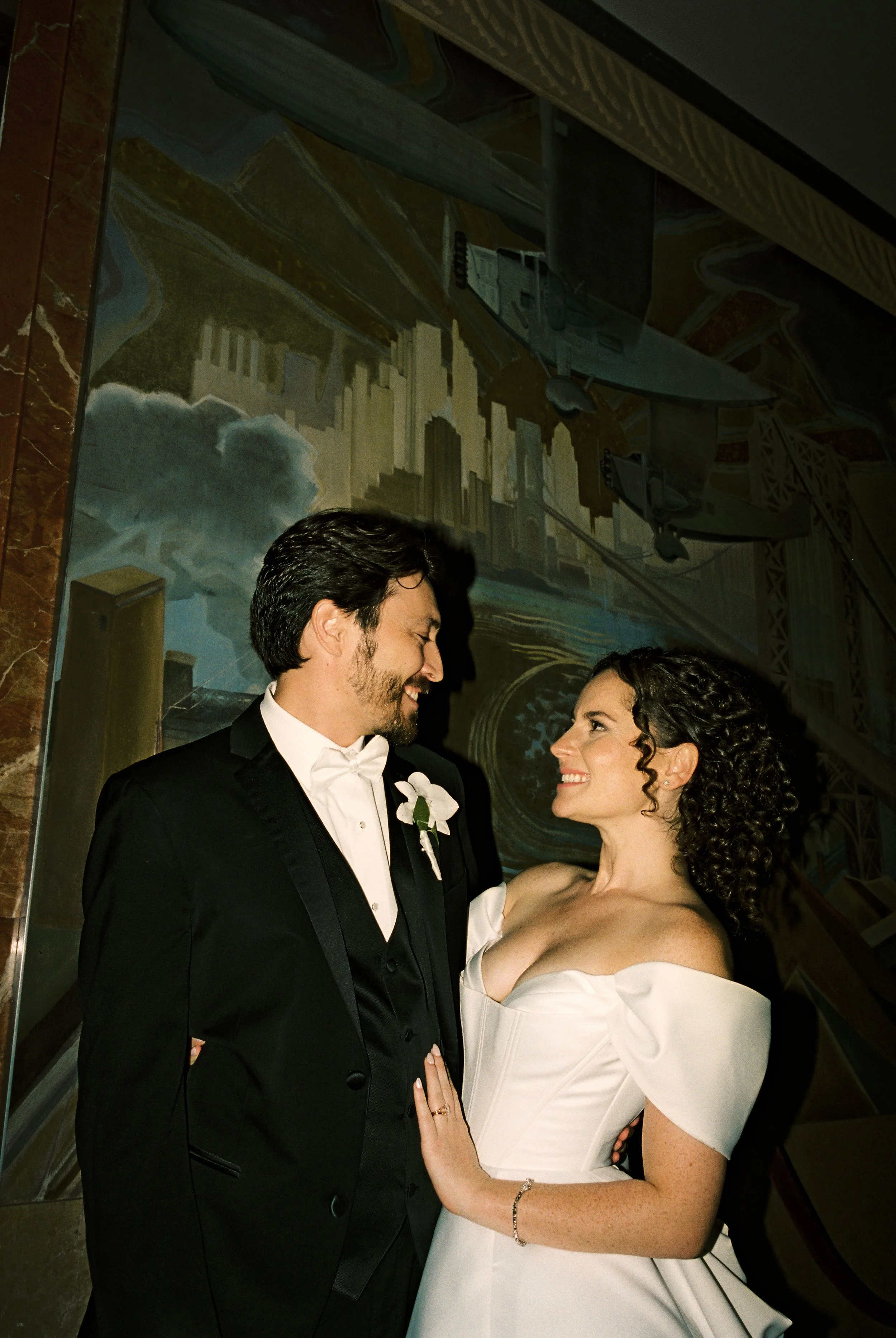Newlywed couple posing together in formal wedding attire, smiling at each other in front of a large aviation-themed mural.