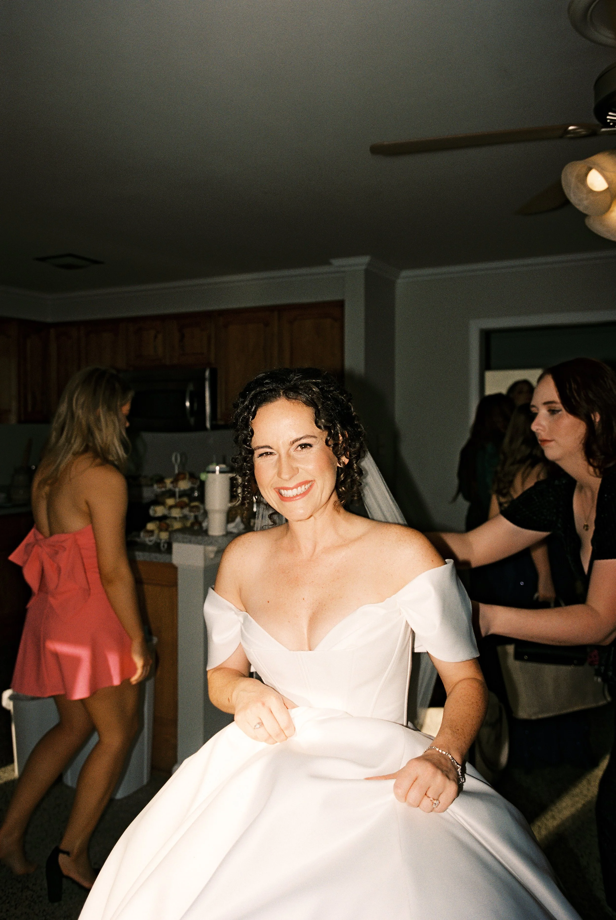 The bride smiles brightly at the camera while holding up her gown, with guests and late-night celebration activity visible in the background.