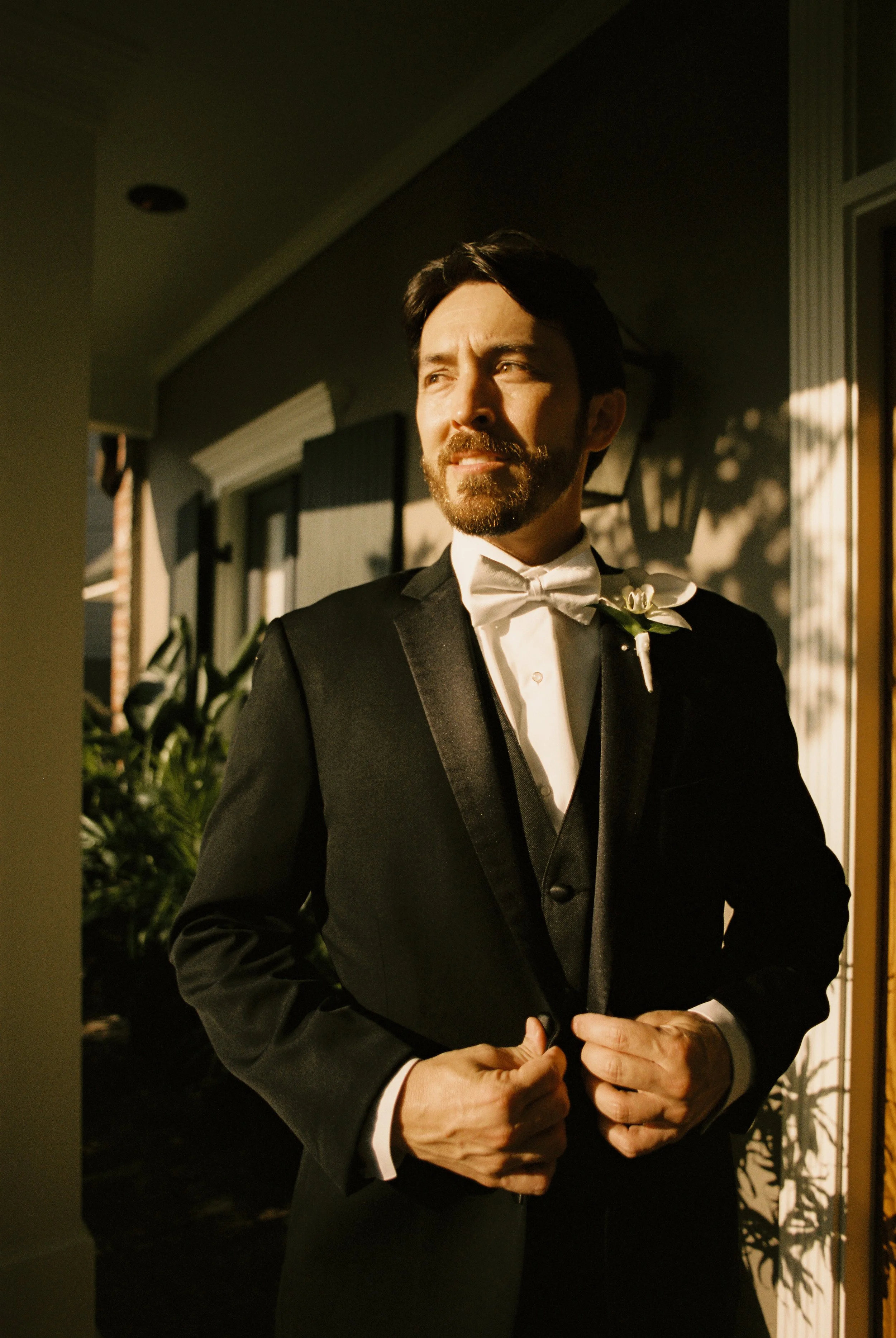 A cinematic portrait of the groom in formal black tie attire, standing in warm golden light with a confident, reflective expression.