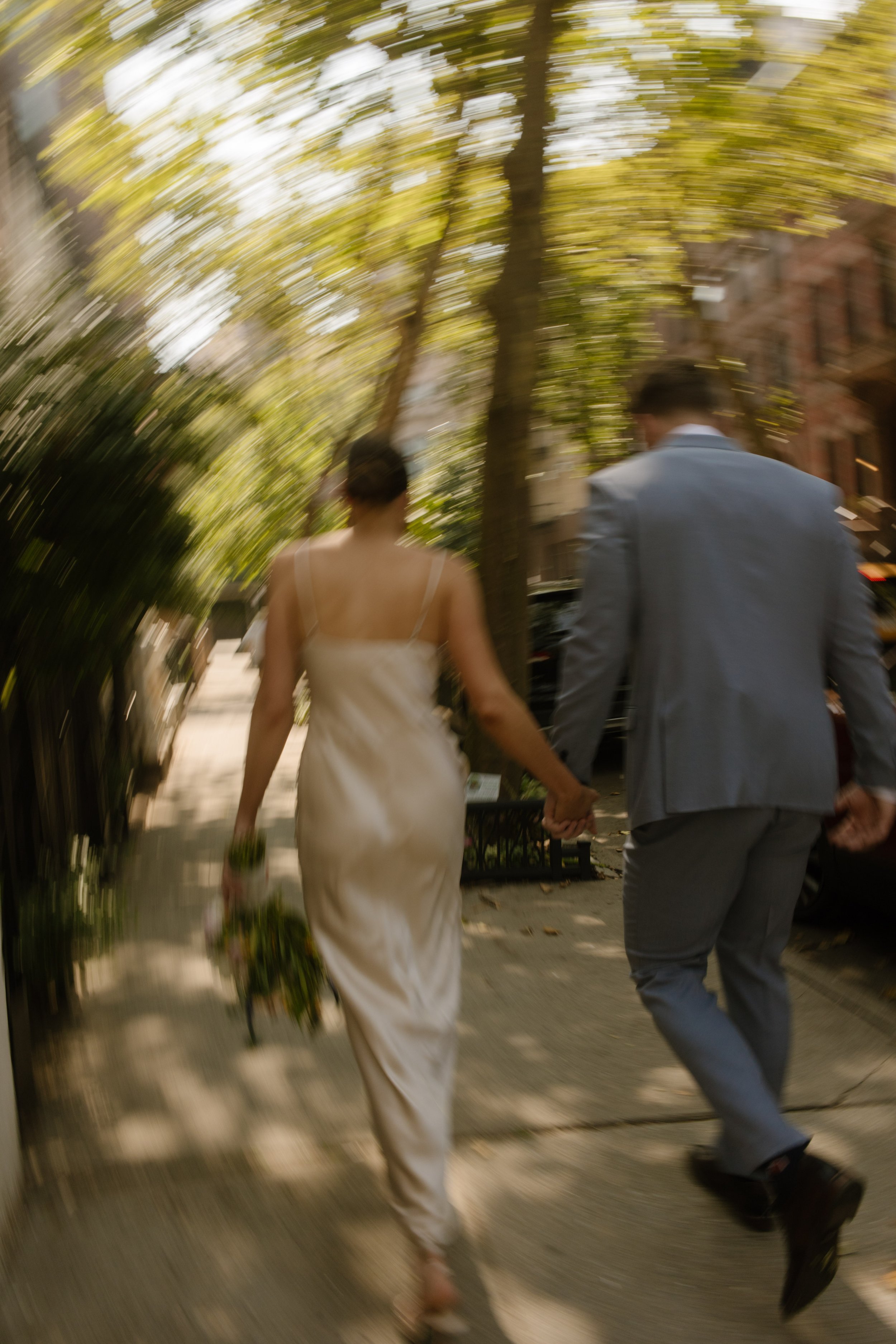 Bride and groom walking hand in hand down a tree-lined sidewalk with artistic motion blur after their wedding.