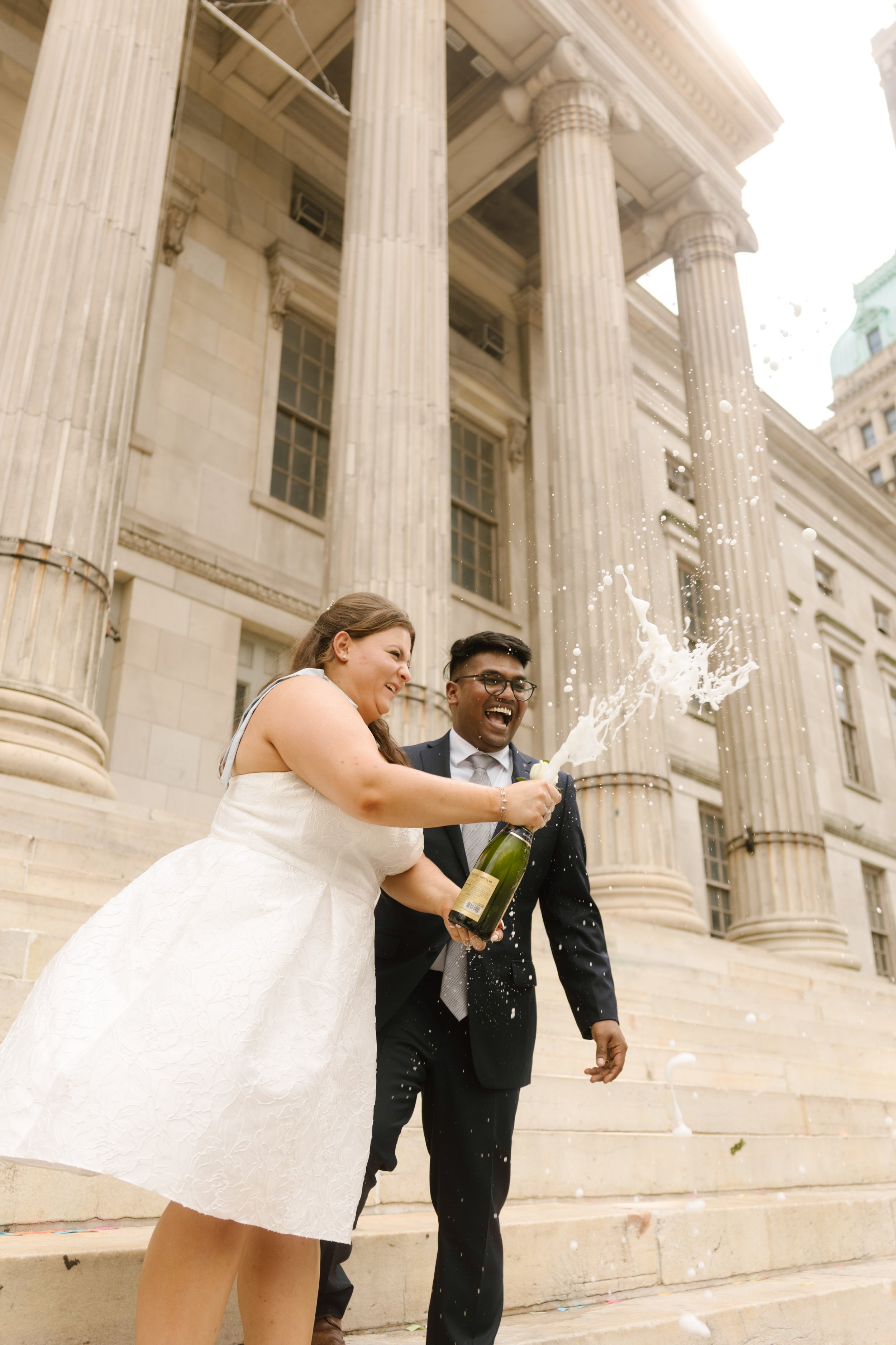 Bride and groom celebrating with a champagne spray outside a historic courthouse, highlighting unique Wedding Locations in NYC for intimate weddings.