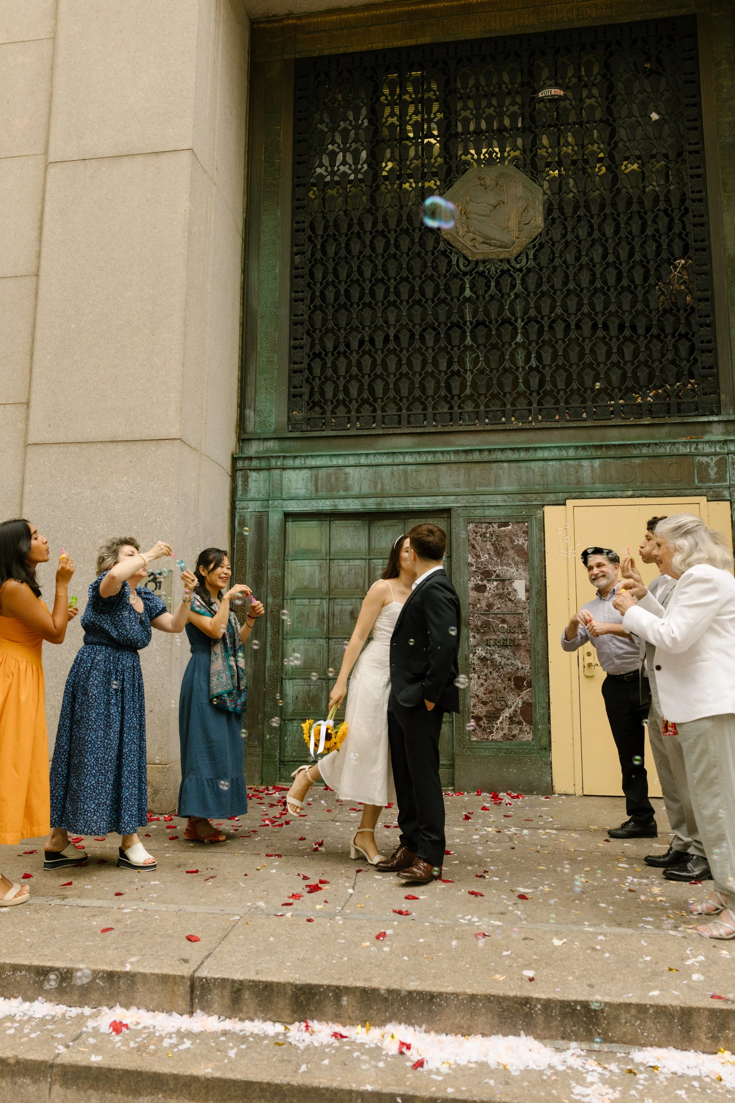 Newlyweds sharing a kiss surrounded by friends and family outside a courthouse after their wedding ceremony.