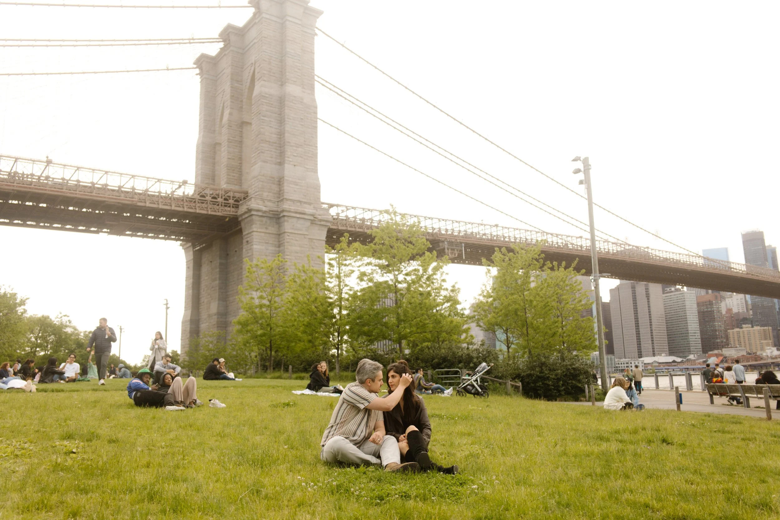 Couple embraces on the lawn beneath the Brooklyn Bridge, highlighting romantic Wedding Locations in NYC.