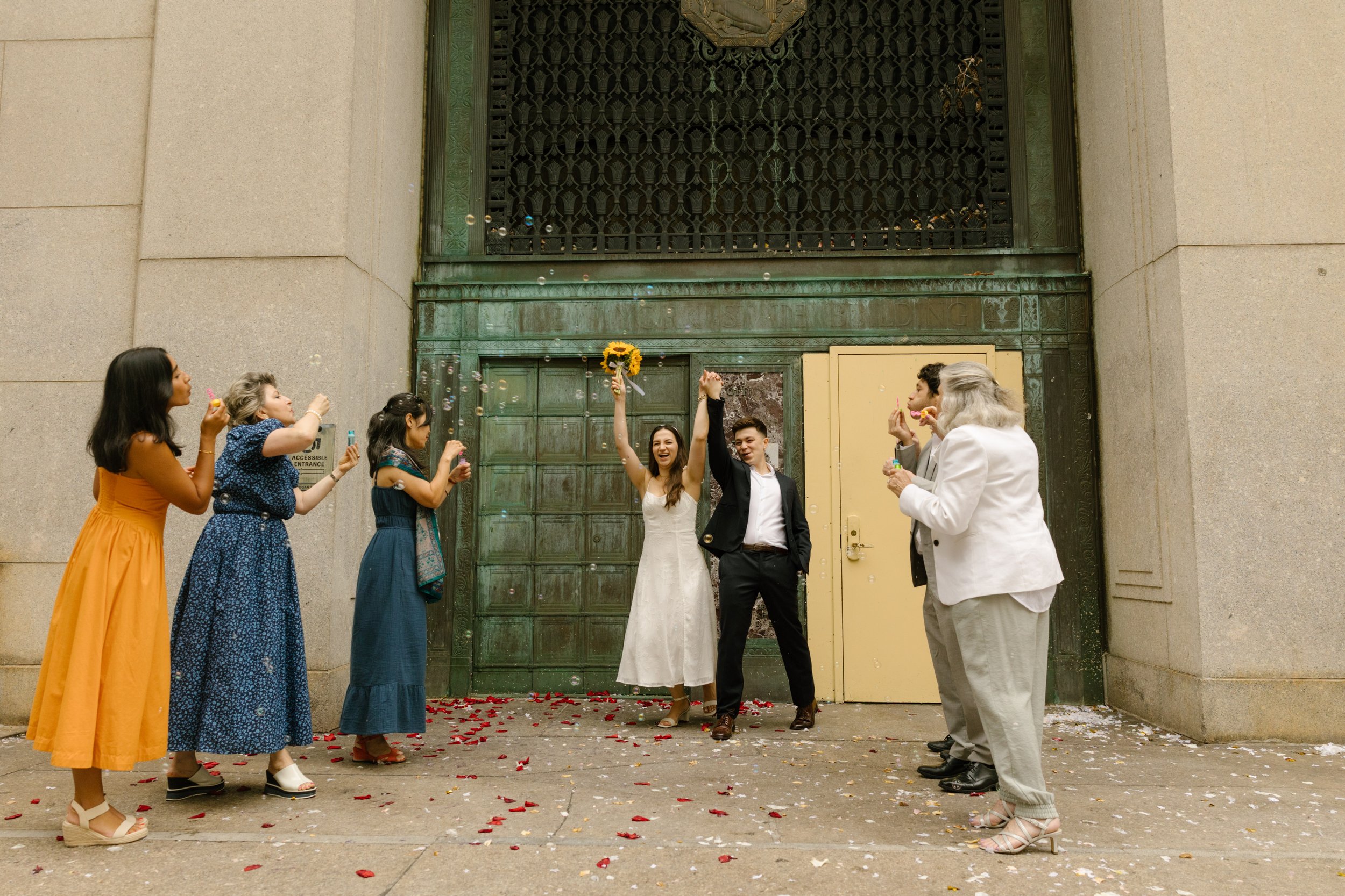 Newlyweds celebrate outside a historic building as friends toss flower petals after their ceremony, featuring joyful Wedding Locations in NYC.