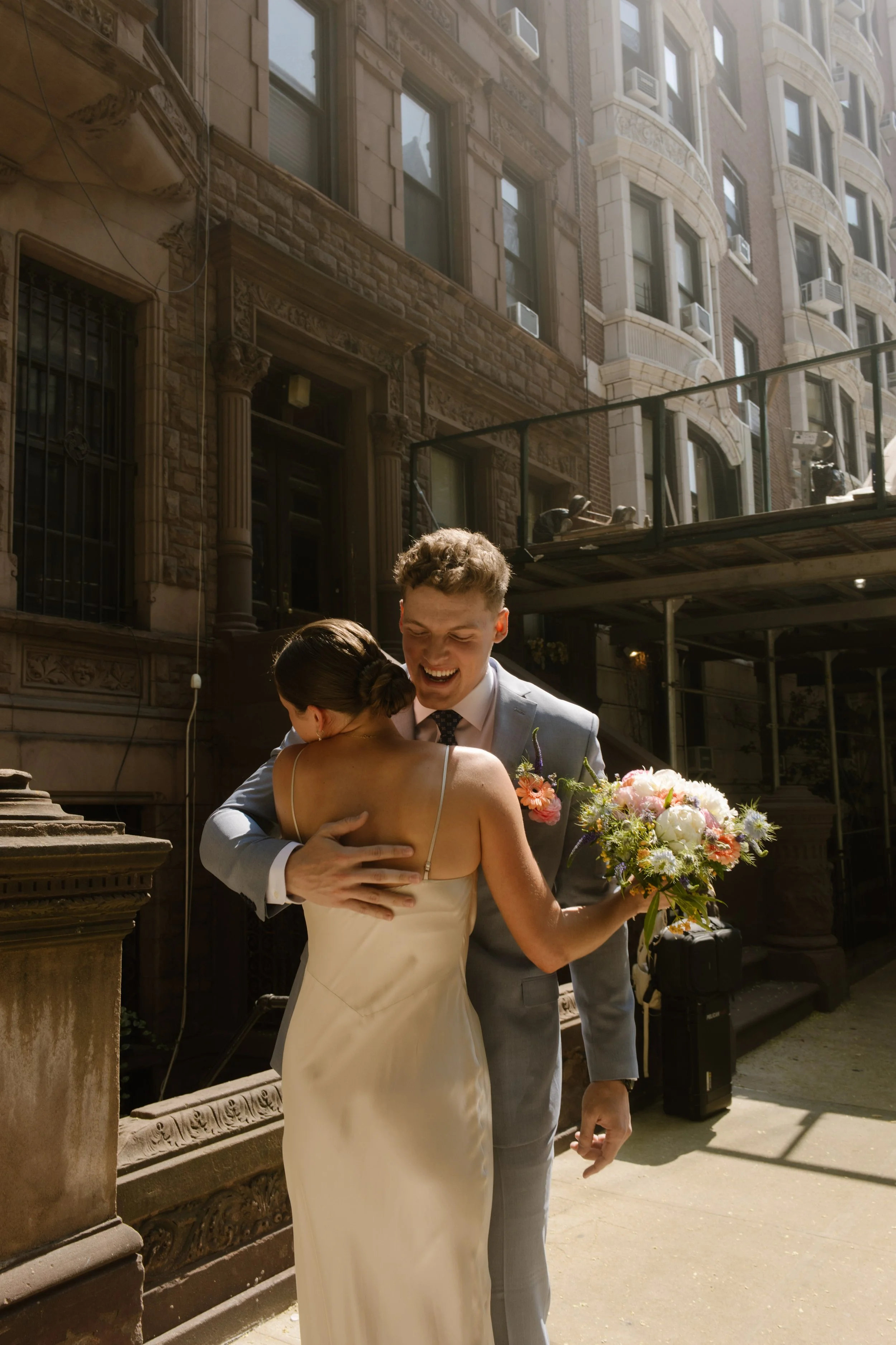Groom hugs the bride outside a historic brownstone with colorful wedding flowers in hand.
