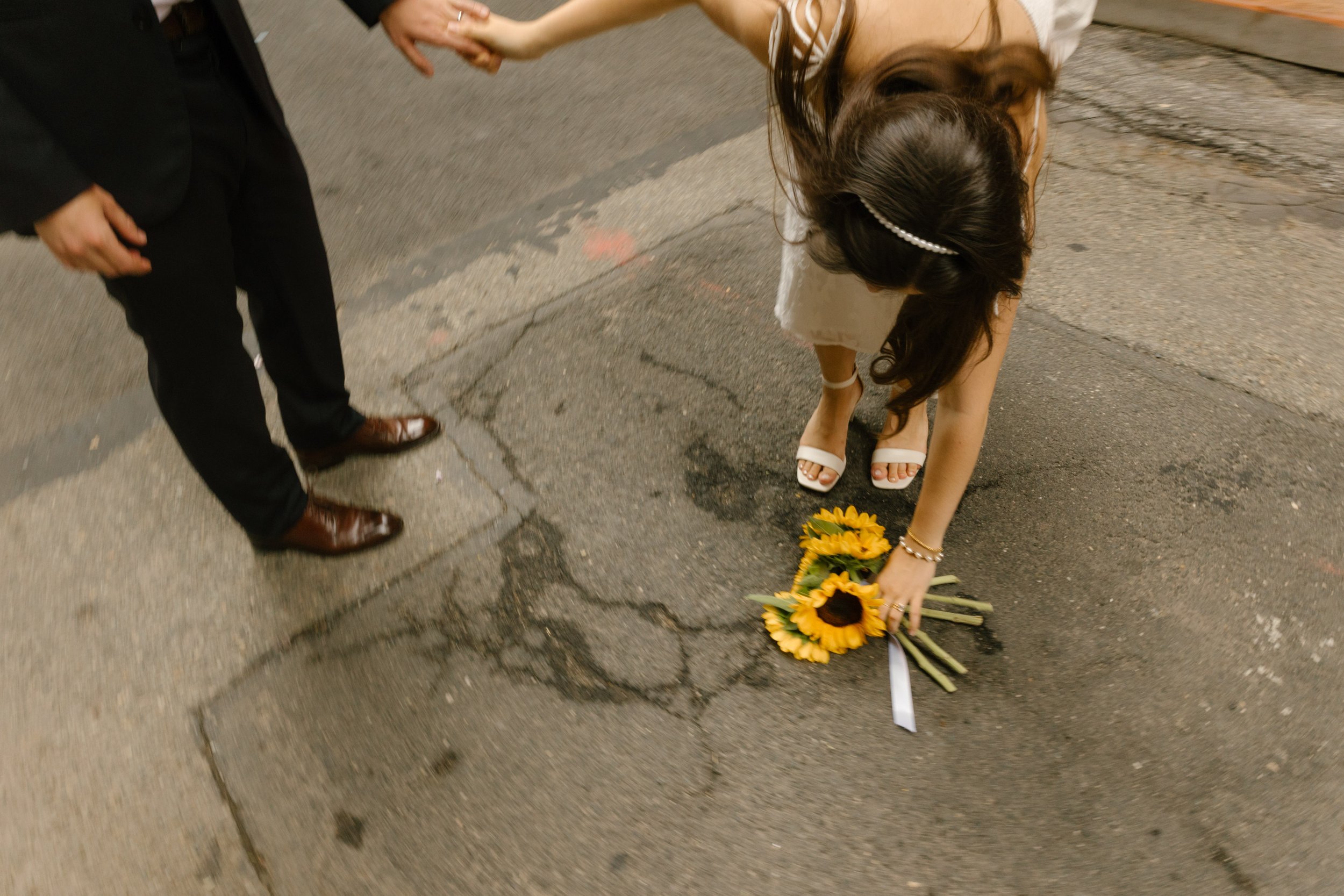 Bride reaches down to pick up a sunflower bouquet after dropping it during a playful city wedding moment.