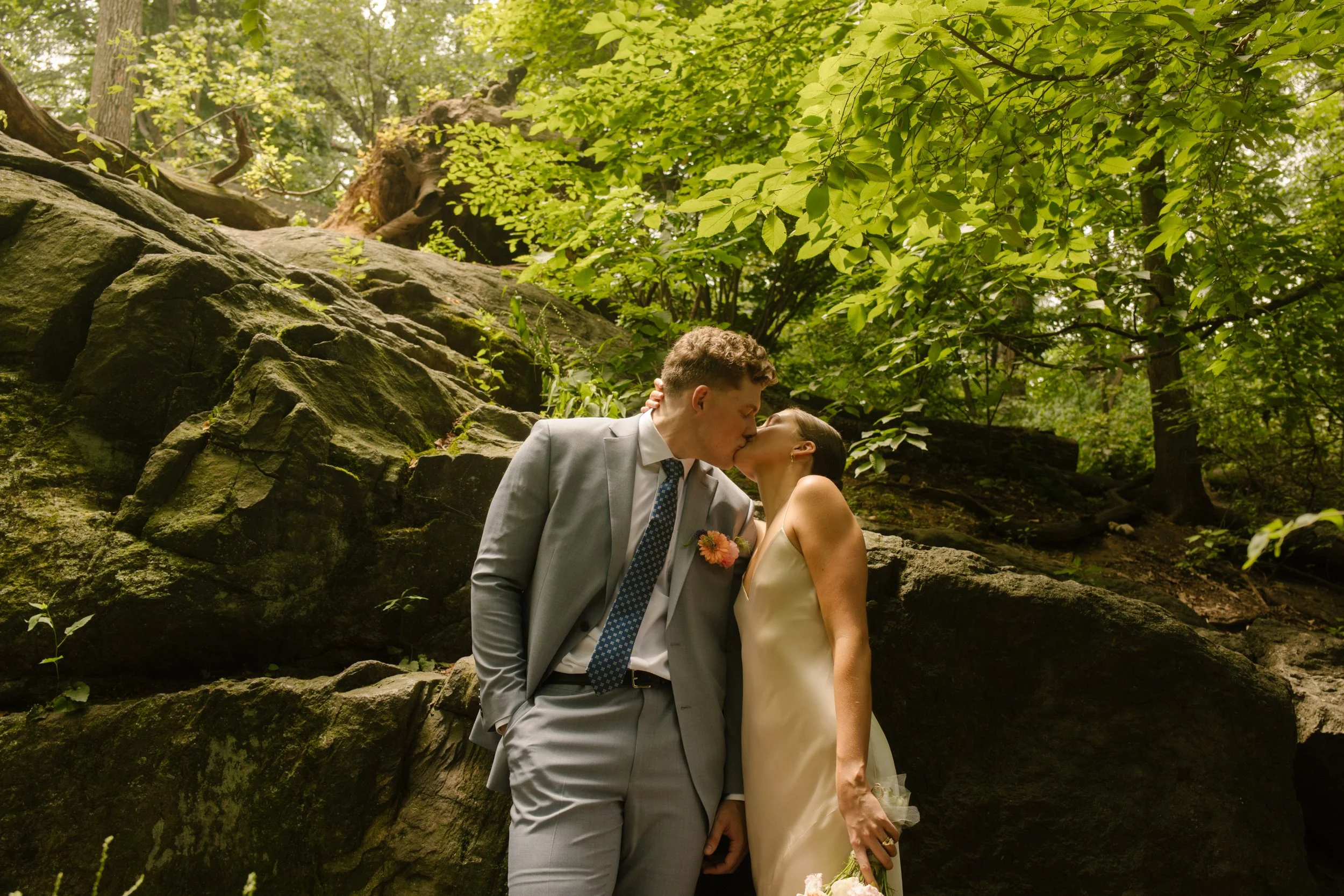 Newlyweds share a kiss beside moss-covered rocks in Central Park, one of the most romantic Wedding Locations in NYC.