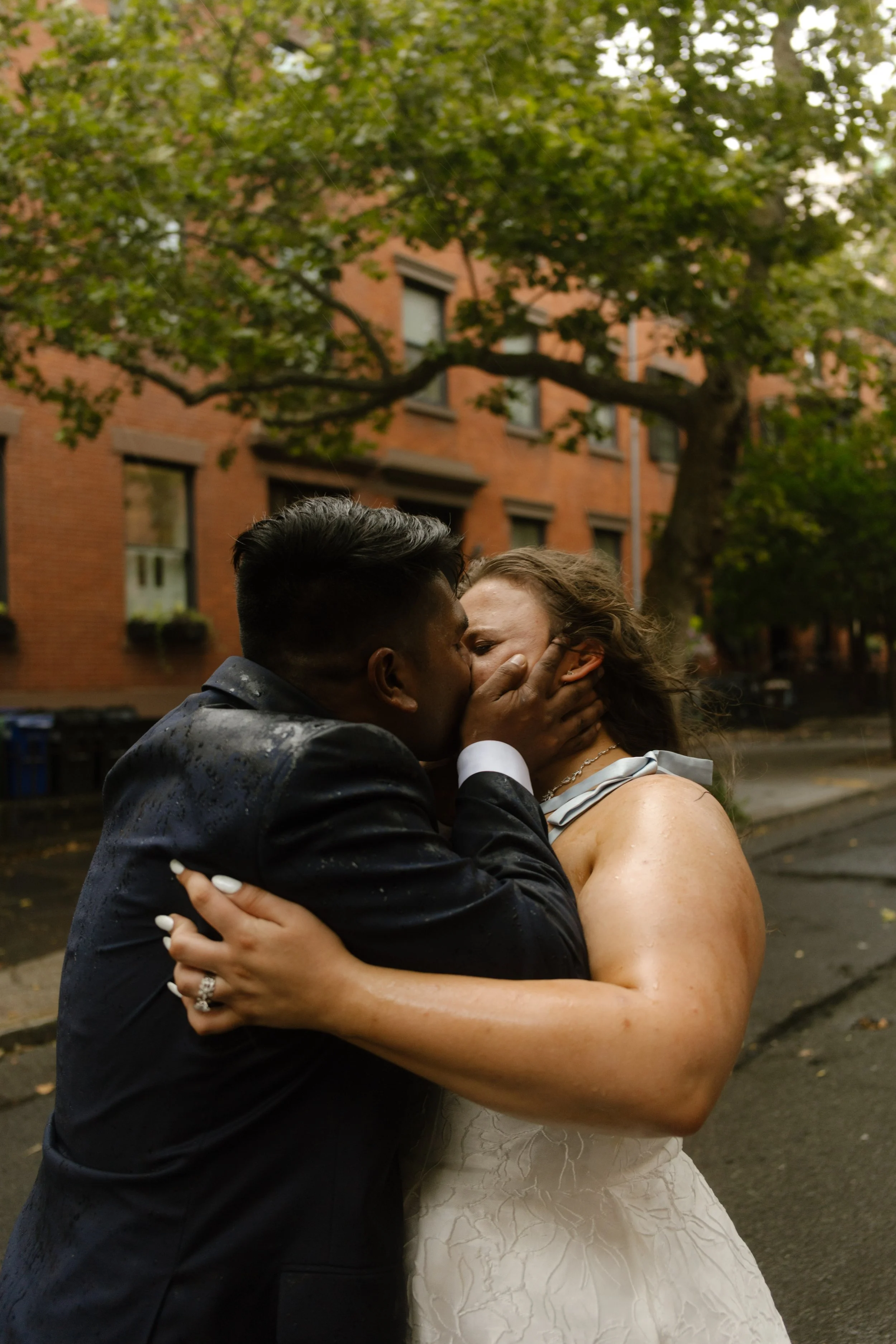 Couple shares a rain-soaked kiss on a tree-lined Brooklyn brownstone street during their wedding portraits.