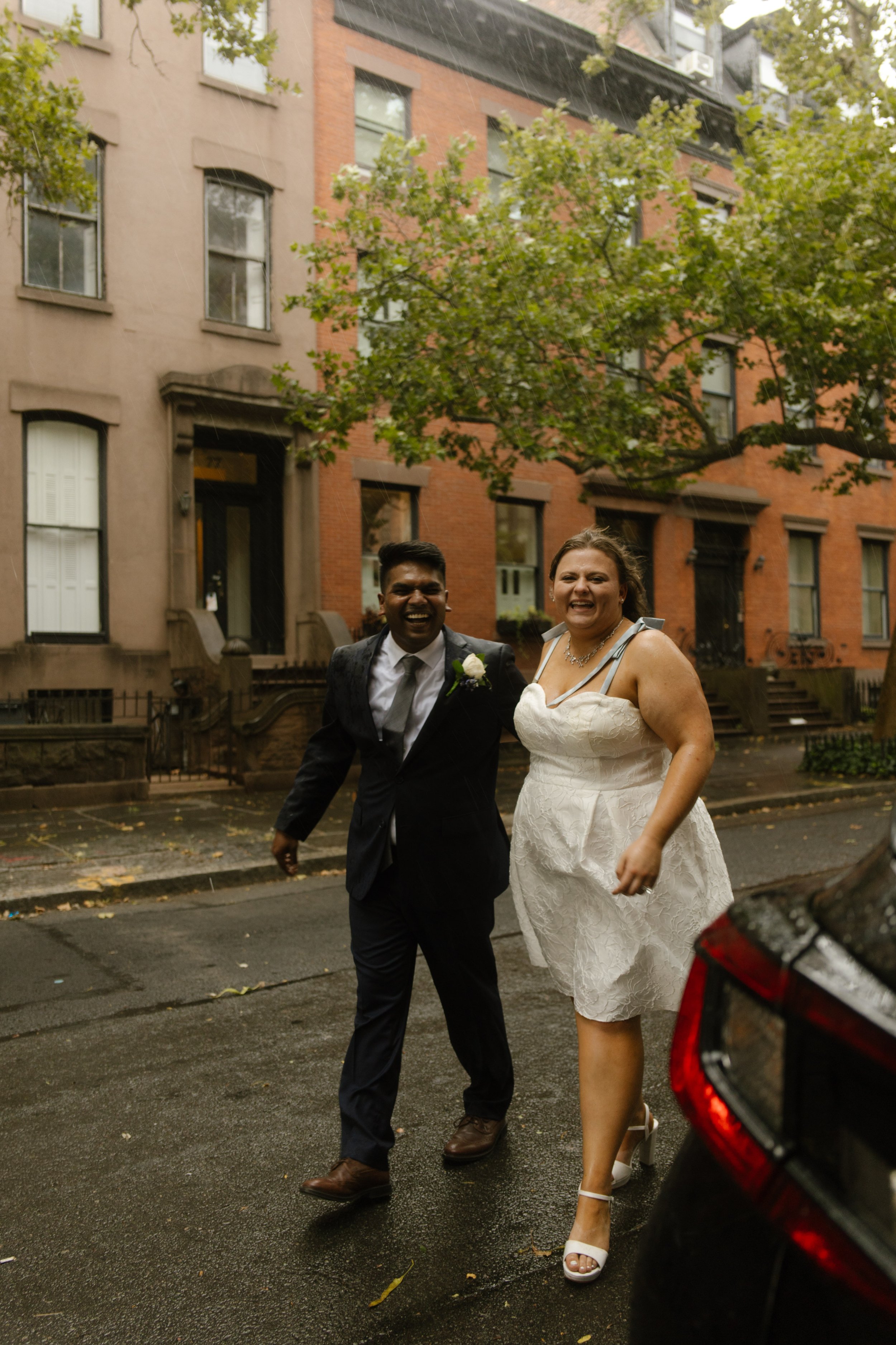 Smiling newlyweds walk hand in hand through a rainy brownstone neighborhood after their intimate city wedding.