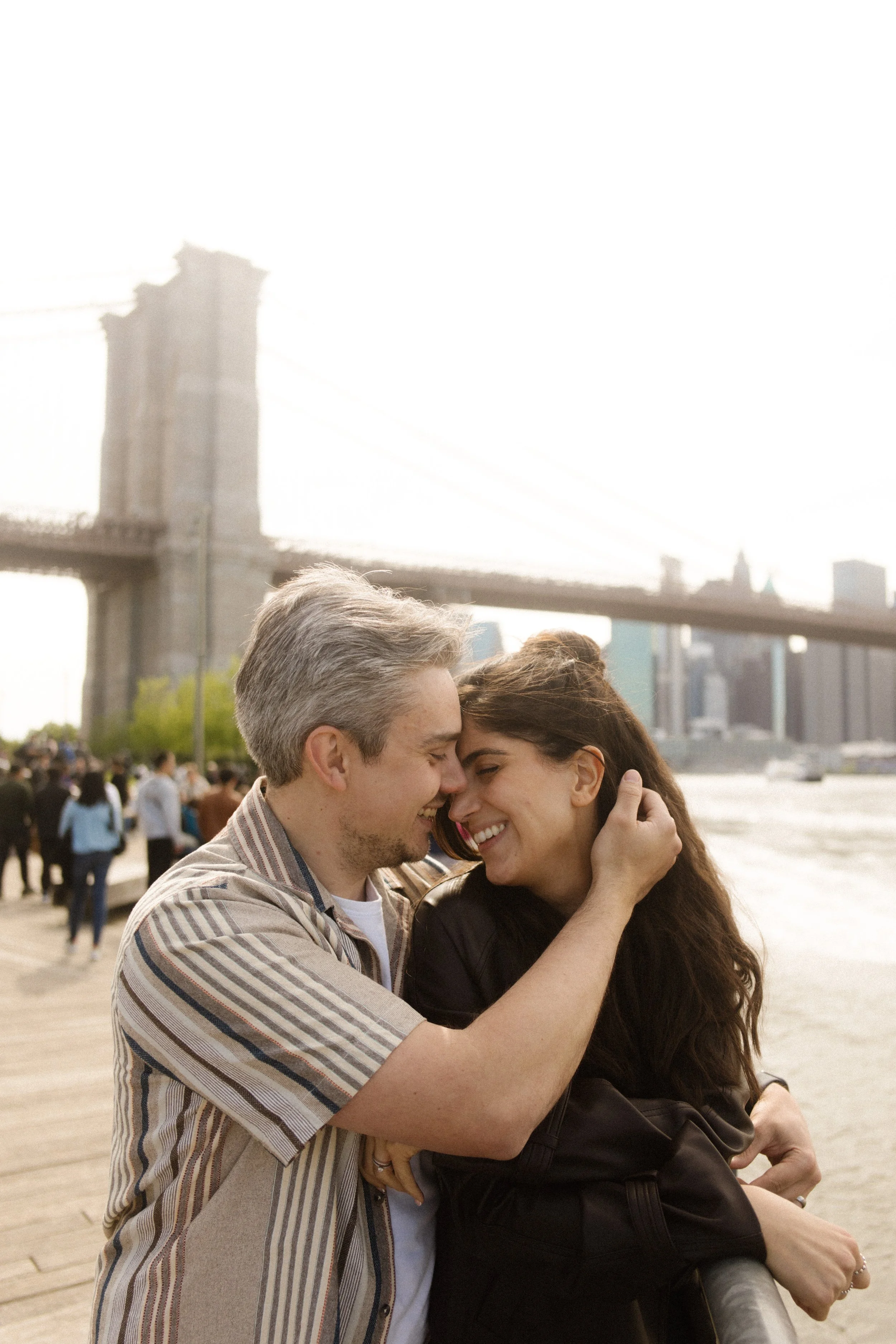 Couple embraces along the Brooklyn waterfront at sunset with the Brooklyn Bridge behind them, highlighting one of the most iconic Wedding Locations in NYC.