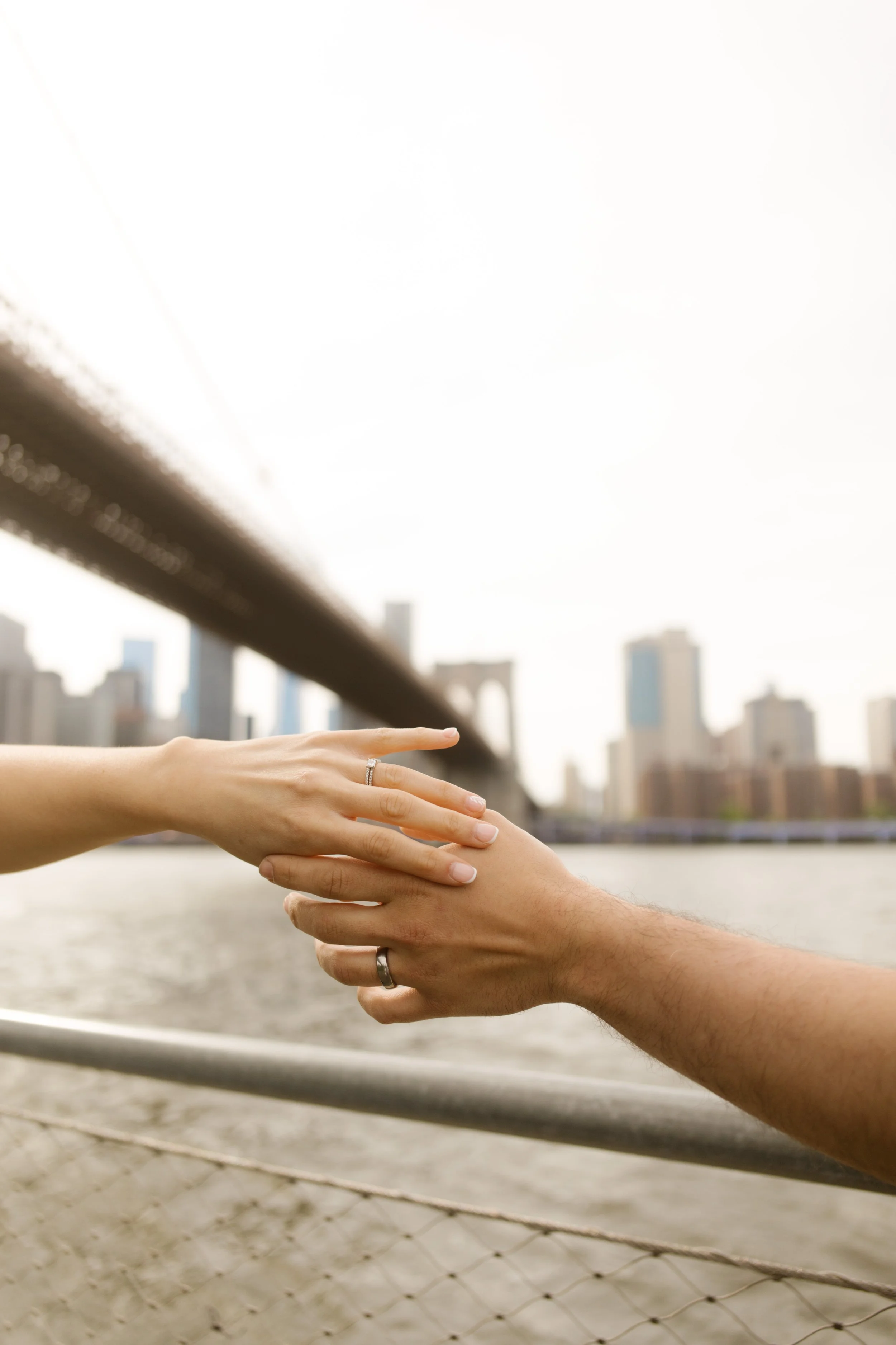 Close-up of wedding bands as a couple reaches for each other's hands with the Brooklyn Bridge and Manhattan skyline in the background, showcasing iconic Wedding Locations in NYC.
