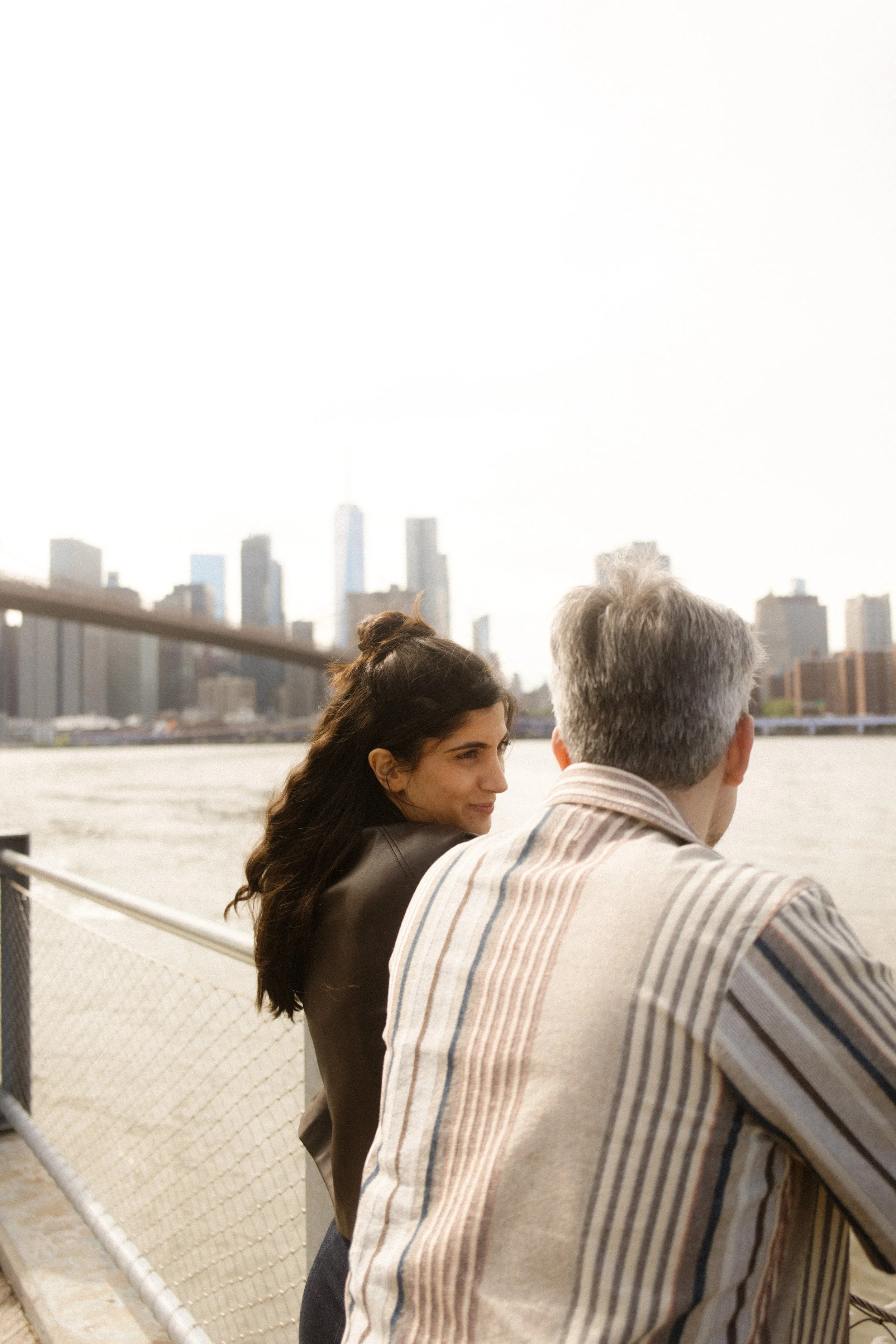 Couple looks out over the East River and Manhattan skyline beneath the Brooklyn Bridge during a romantic engagement session.