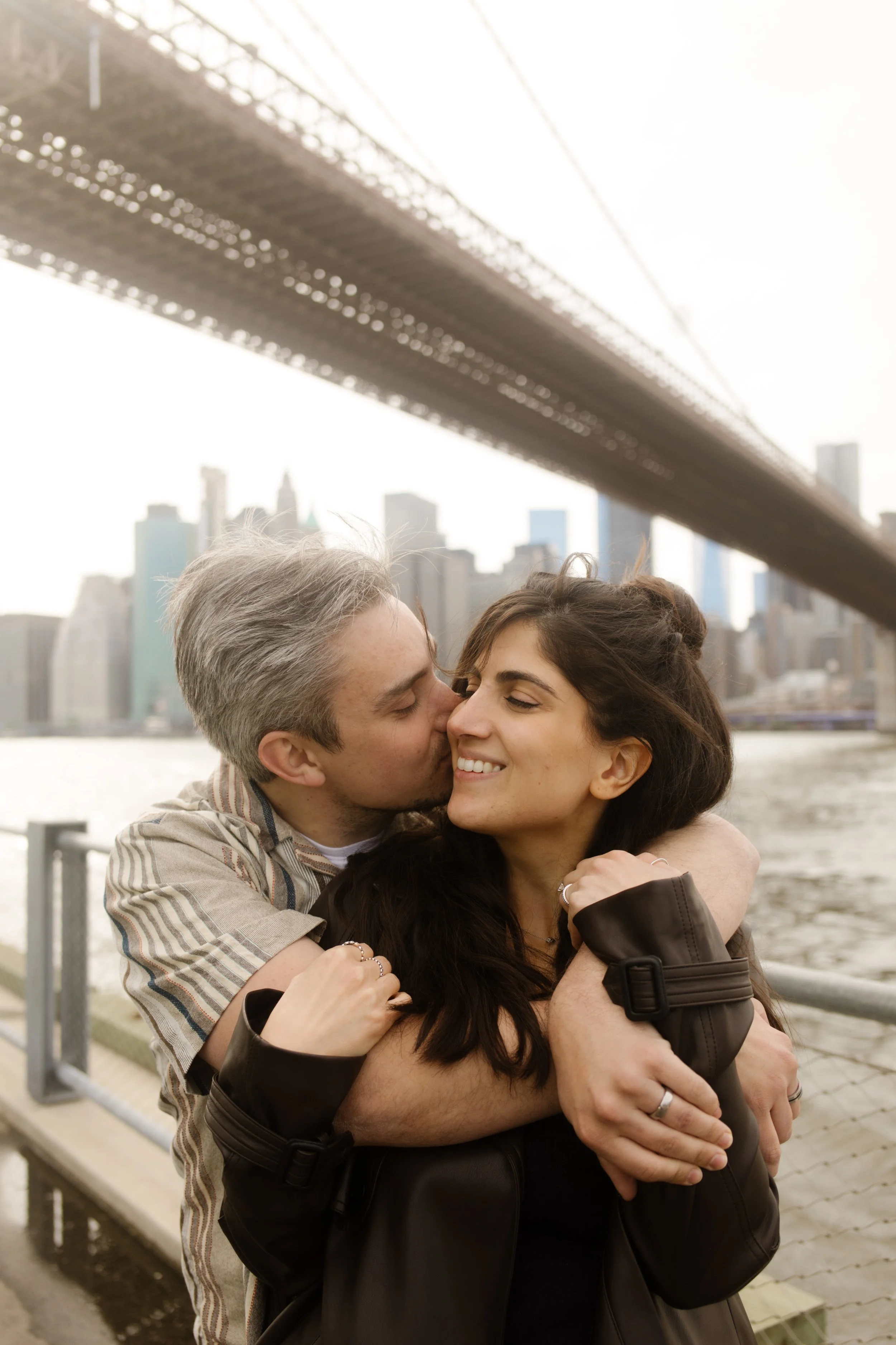 Groom hugs his partner from behind as they smile beneath the Brooklyn Bridge at one of the most romantic Wedding Locations in NYC.