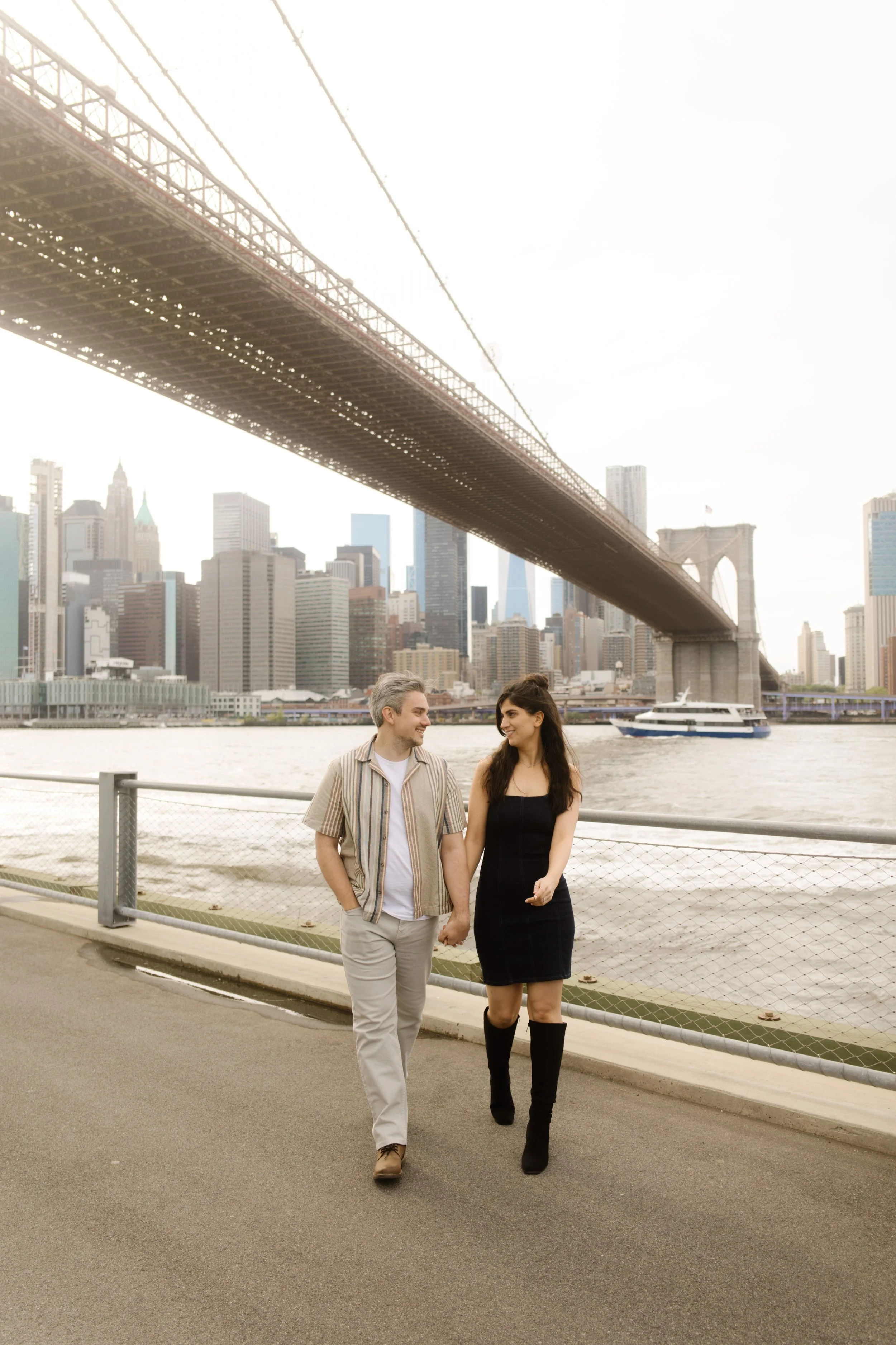 Couple walks hand in hand along the Brooklyn waterfront with sweeping views of the Brooklyn Bridge and Lower Manhattan.