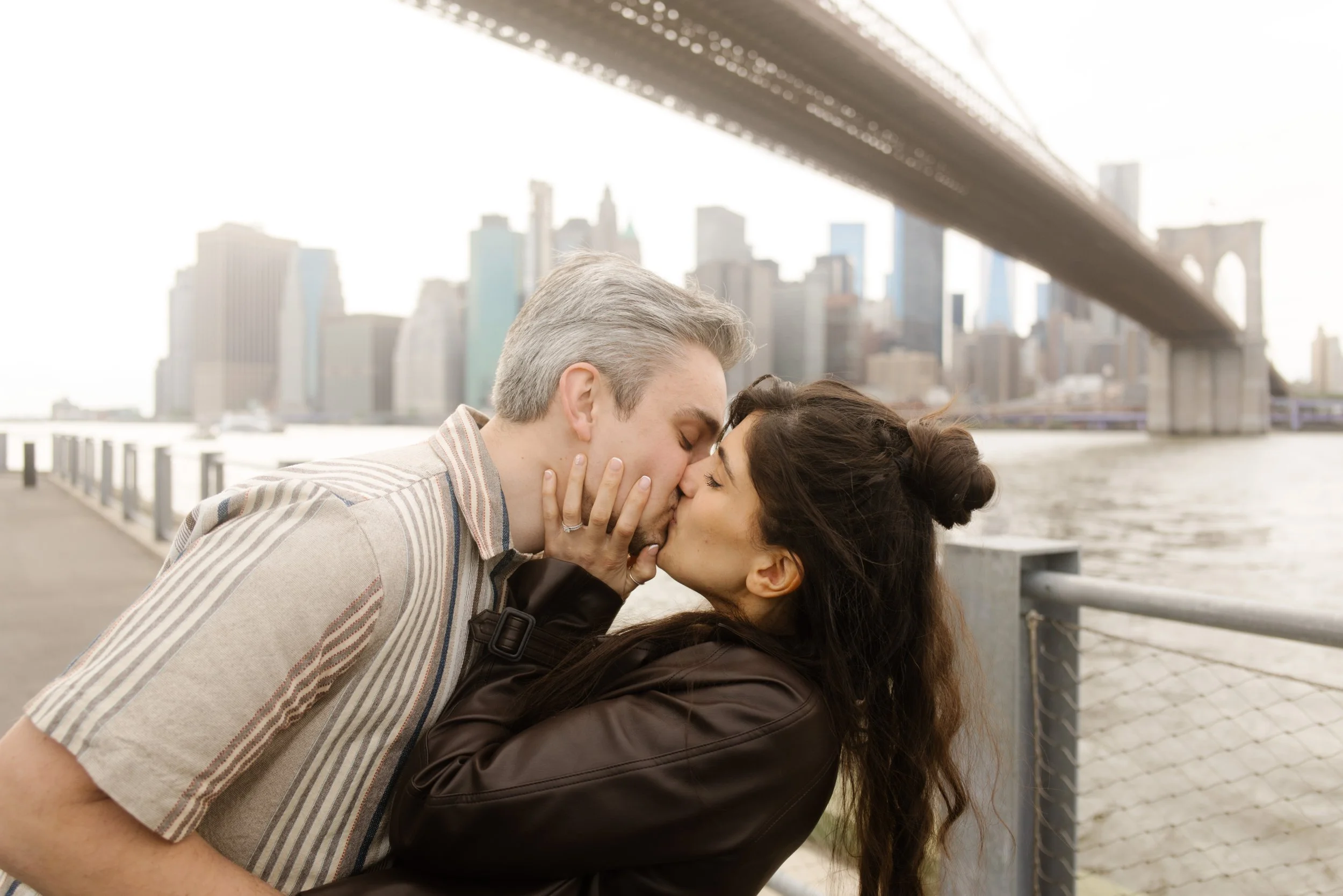 Couple shares a kiss along the Brooklyn waterfront with the Brooklyn Bridge and city skyline creating a classic New York backdrop.