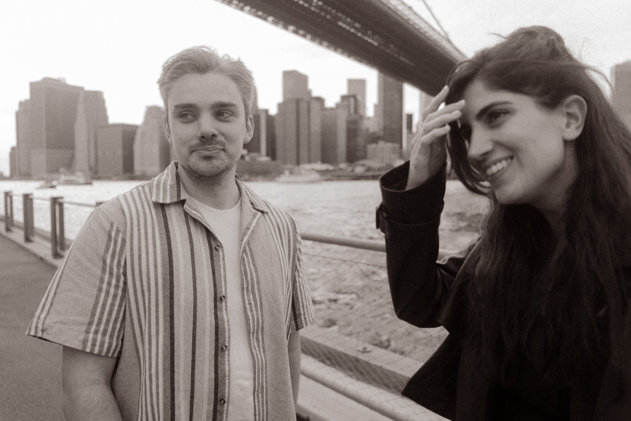 Candid black-and-white portrait of a smiling couple along the Brooklyn waterfront beneath the Brooklyn Bridge.