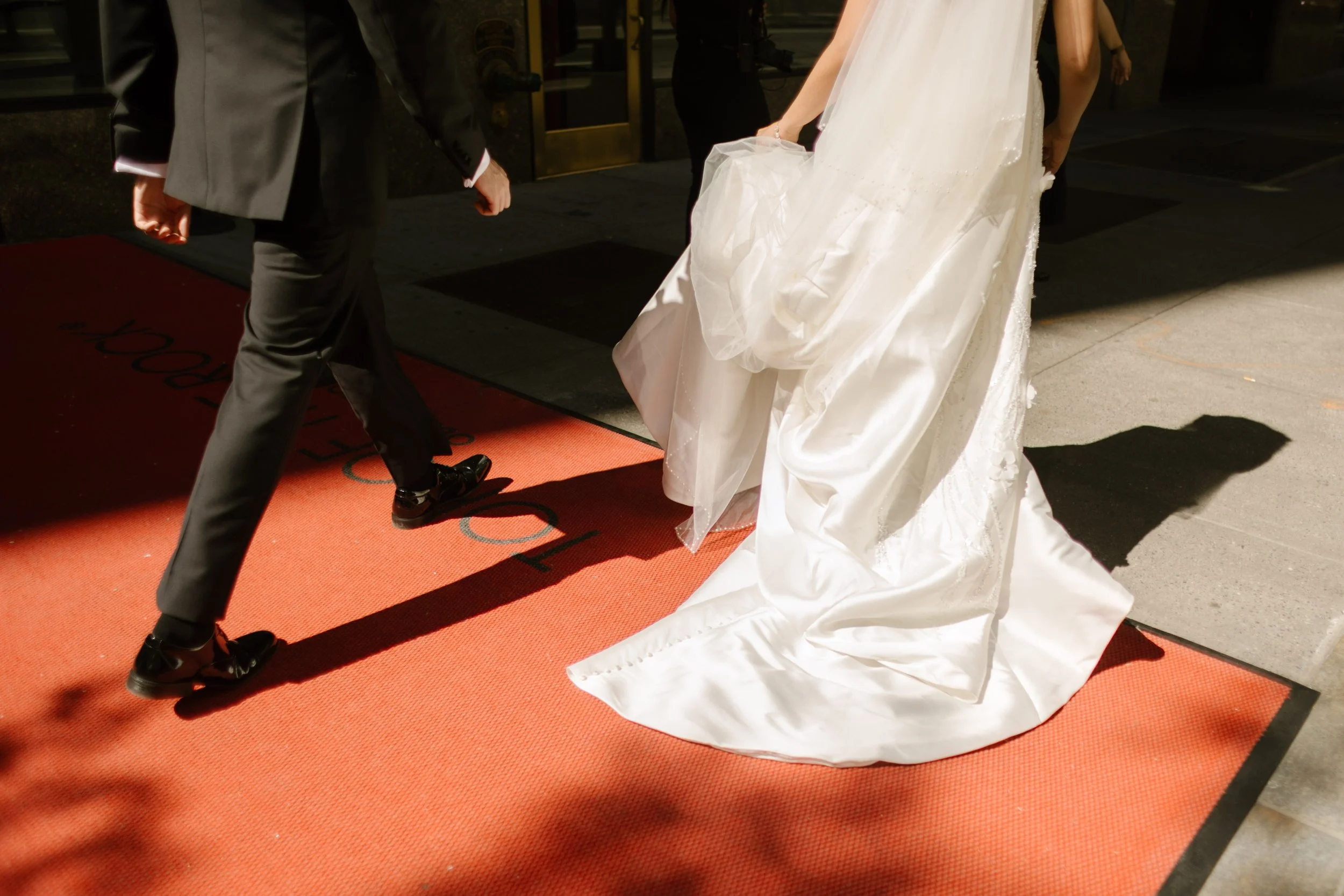 Bride gathers her wedding dress as the couple walks across a hotel entrance with elegant city wedding details.