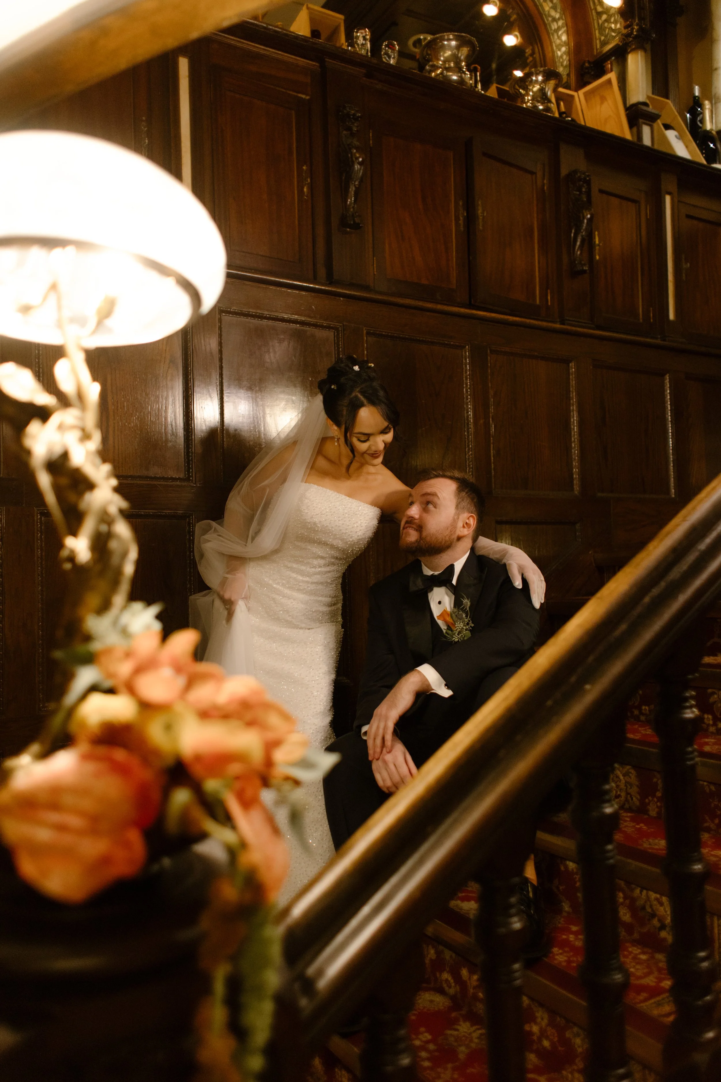 Bride and groom share a quiet moment on a grand historic staircase with warm wood paneling and autumn florals.