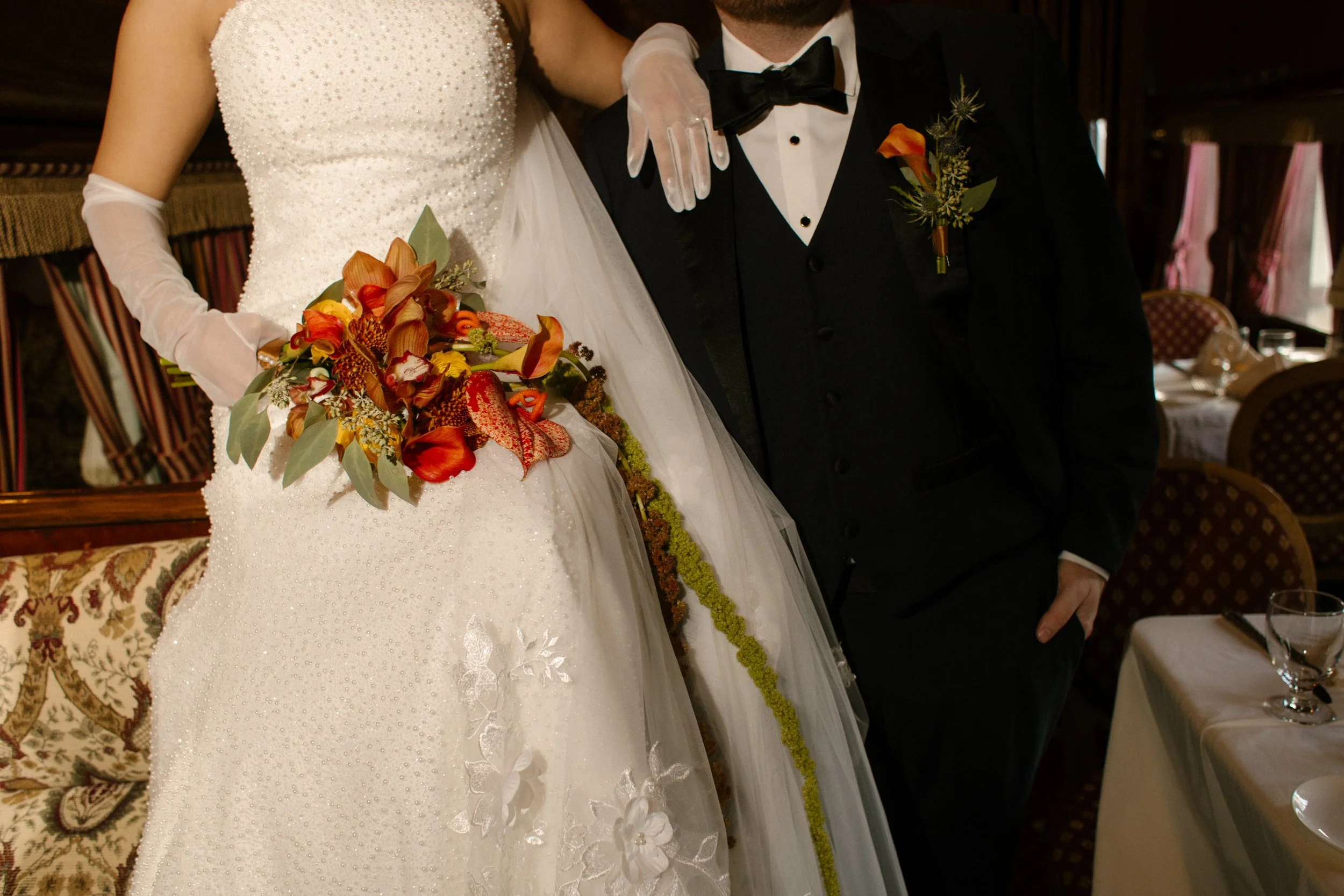 Close-up of the bride's beaded gown and cascading autumn bouquet beside the groom's classic black tuxedo.