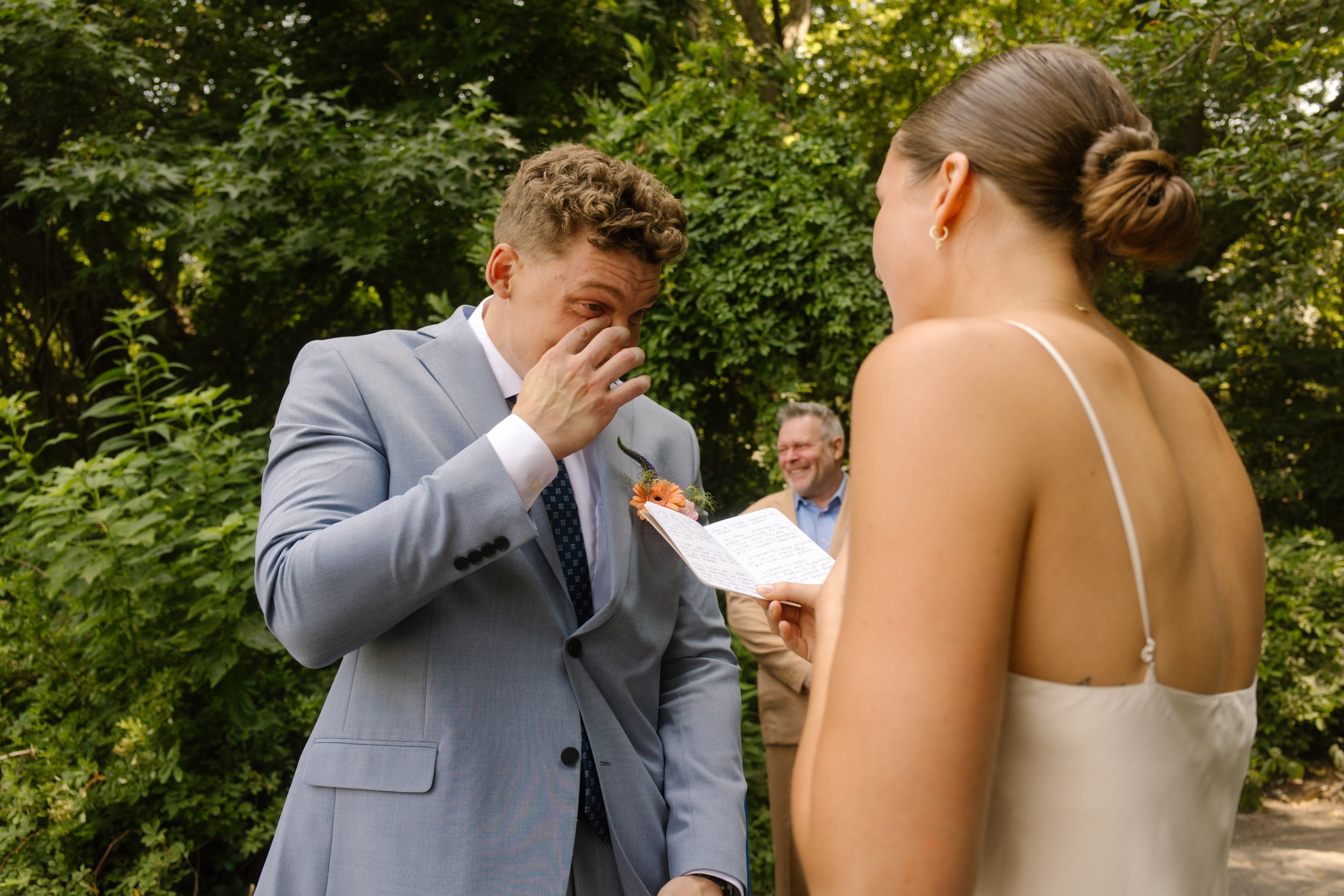 Groom wipes away tears as the bride reads handwritten vows during their emotional outdoor wedding ceremony.